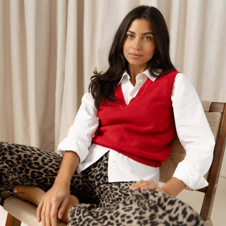 A model lounging in a wooden armchair against a beige curtain wearing a white button-up shirt under a red cashmere sweater vest with a V neckline and leopard print jeans