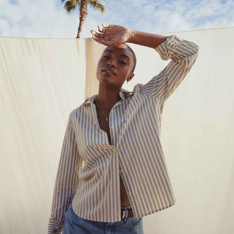 A model standing in front of a white linen sheet that hangs on a clothing line under a sunny sky with a palm tree appearing from the top. The model wears a blue and white striped shirt, left undone at the neckline and the hem. The shirt is the Anneliese shirt from Saint and Sofia.