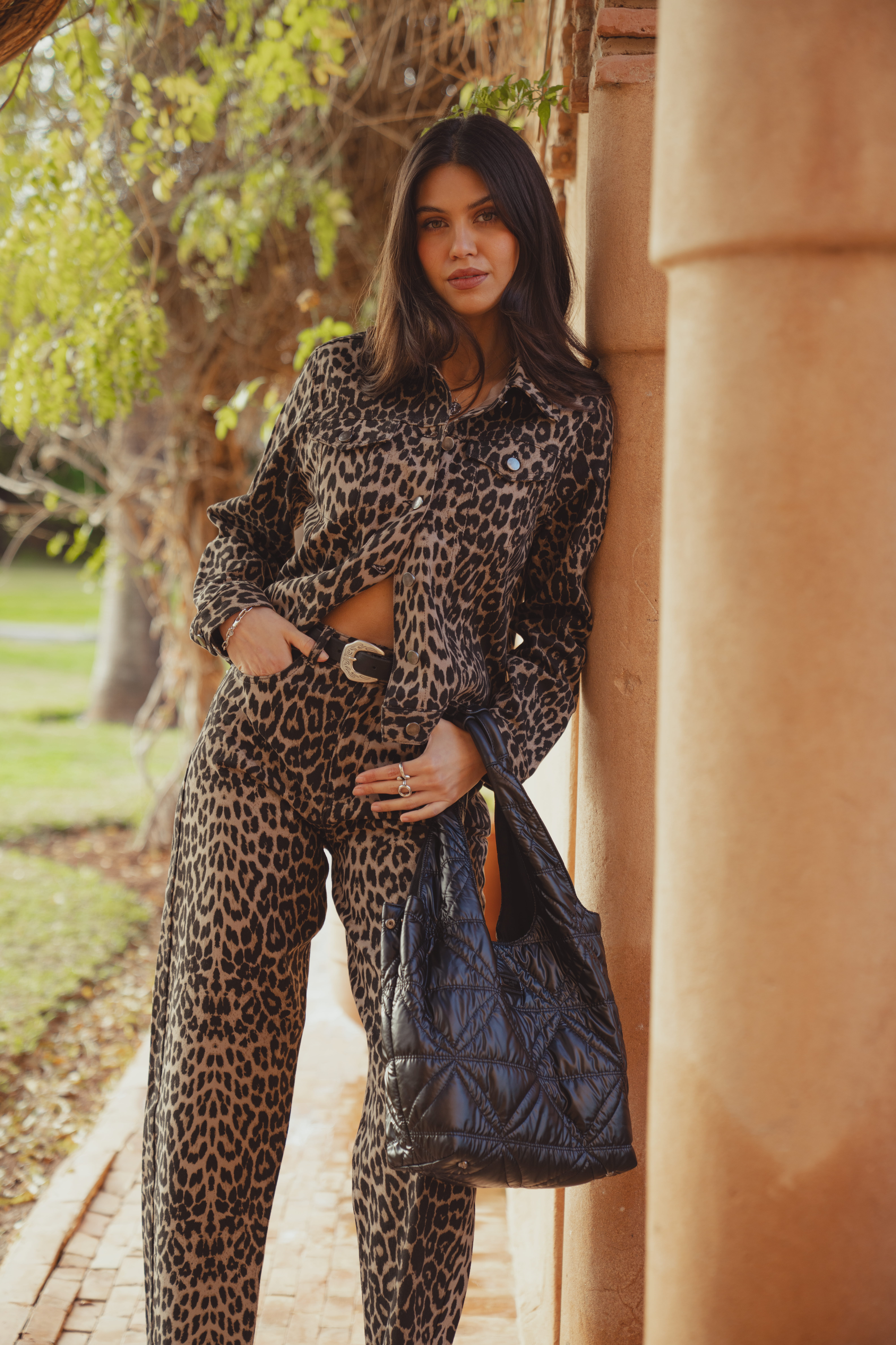 A model standing outside leaning against a pillar wearing the Saint and Sofia Bowie Denim Jacket in leopard print with the matching Barrel Leg Jeans in leopard. The look is styled with a leather belt and a quilted nylon tote bag.