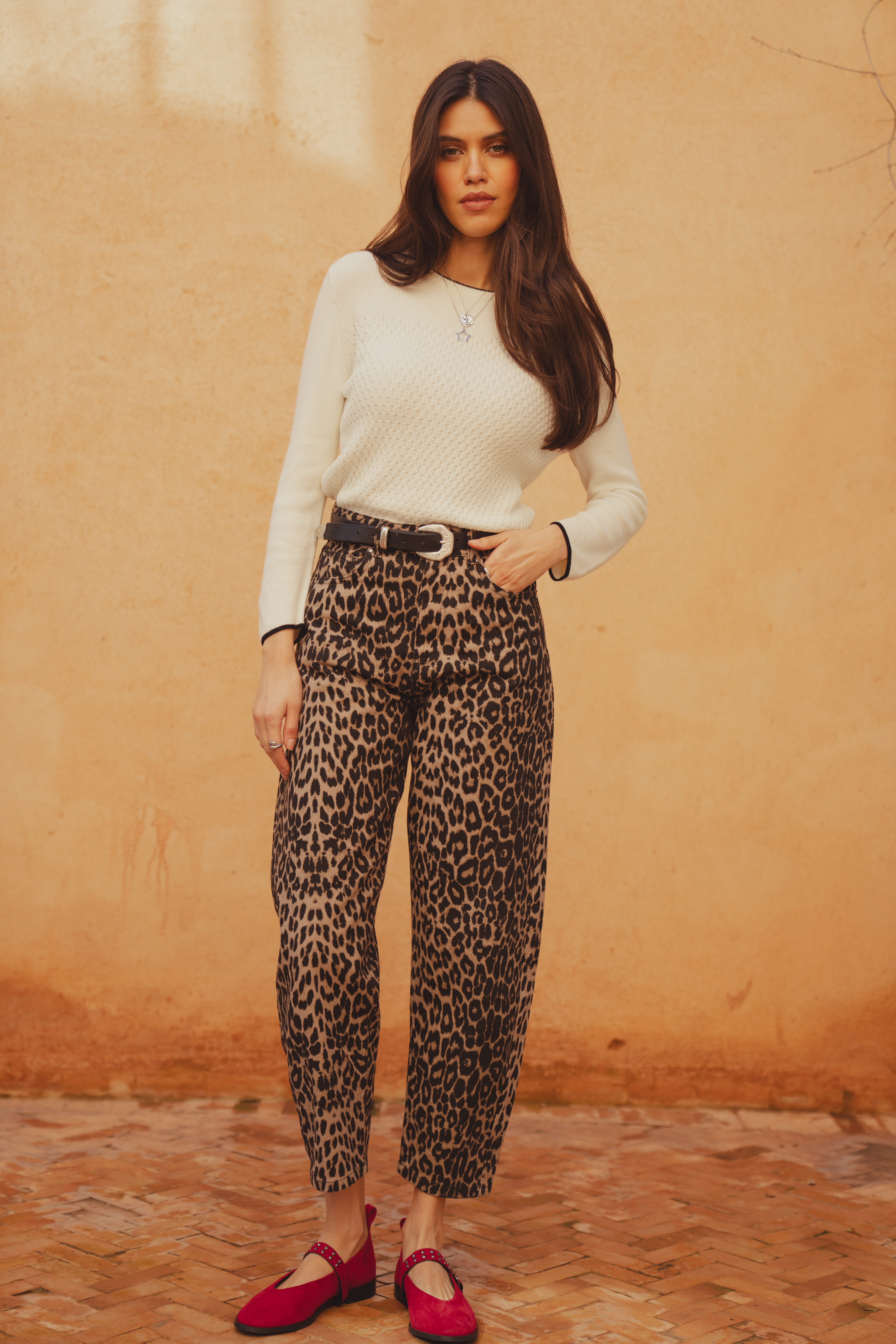 A model standing in front of a clay wall, looking into the camera. She wears the Saint and Sofia Honeycomb Rib Jumper in Cream. It features a diamond knit texture with contrasting trims at the hem and the sleeves. She wears it with the Barrel Leg Jeans in leoapard and styles it with a black leather belt and suede red ballerina flats.