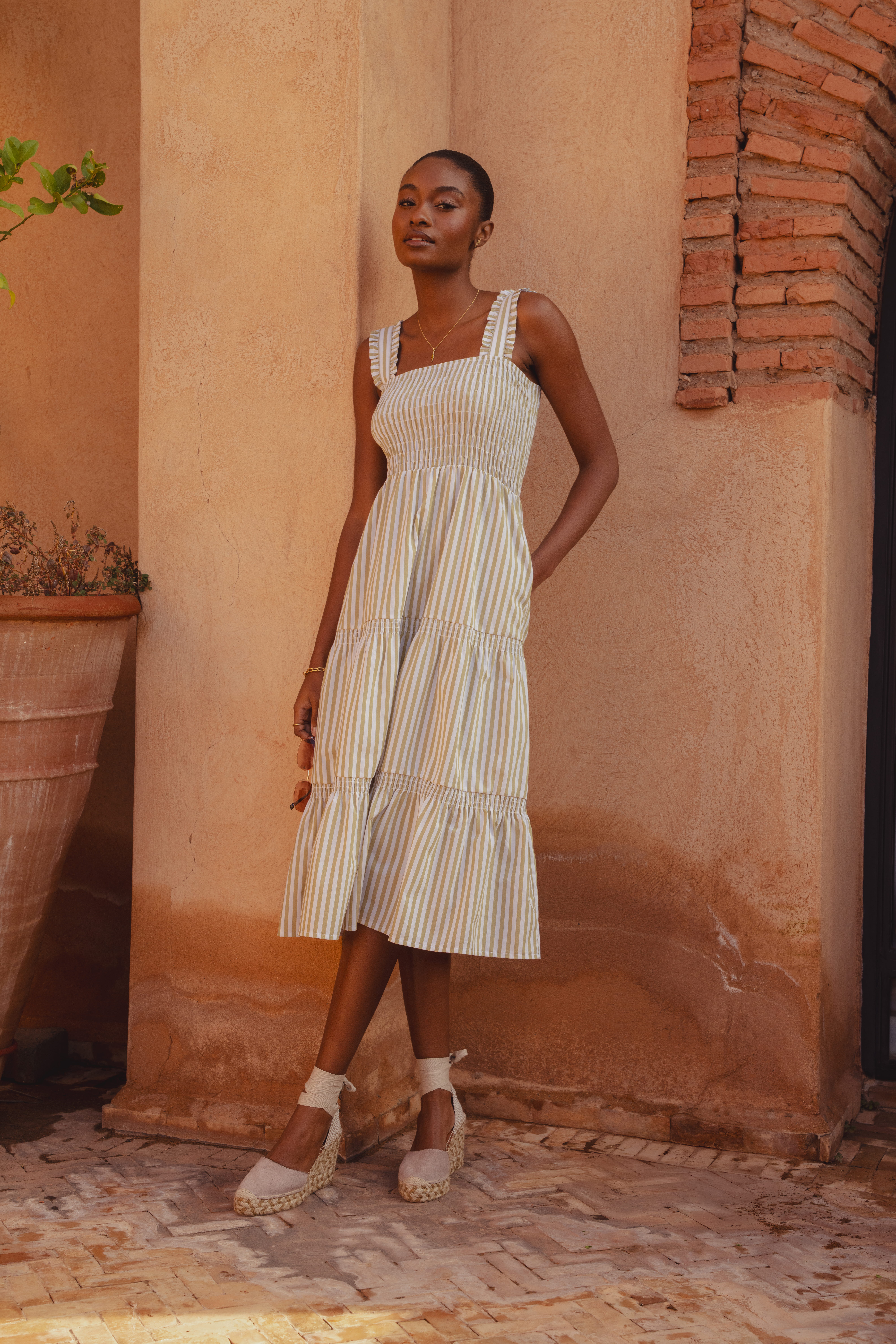 A model standing in front of a clay building wearing the Saint and Sofia Maya Shirring Dress. This sleeveless style features a square neckline, wide straps with frill trim detailing, a shirred bodice, and a tiered midi skirt. It's decorated with green and white stripes for a nautical aesthetic.