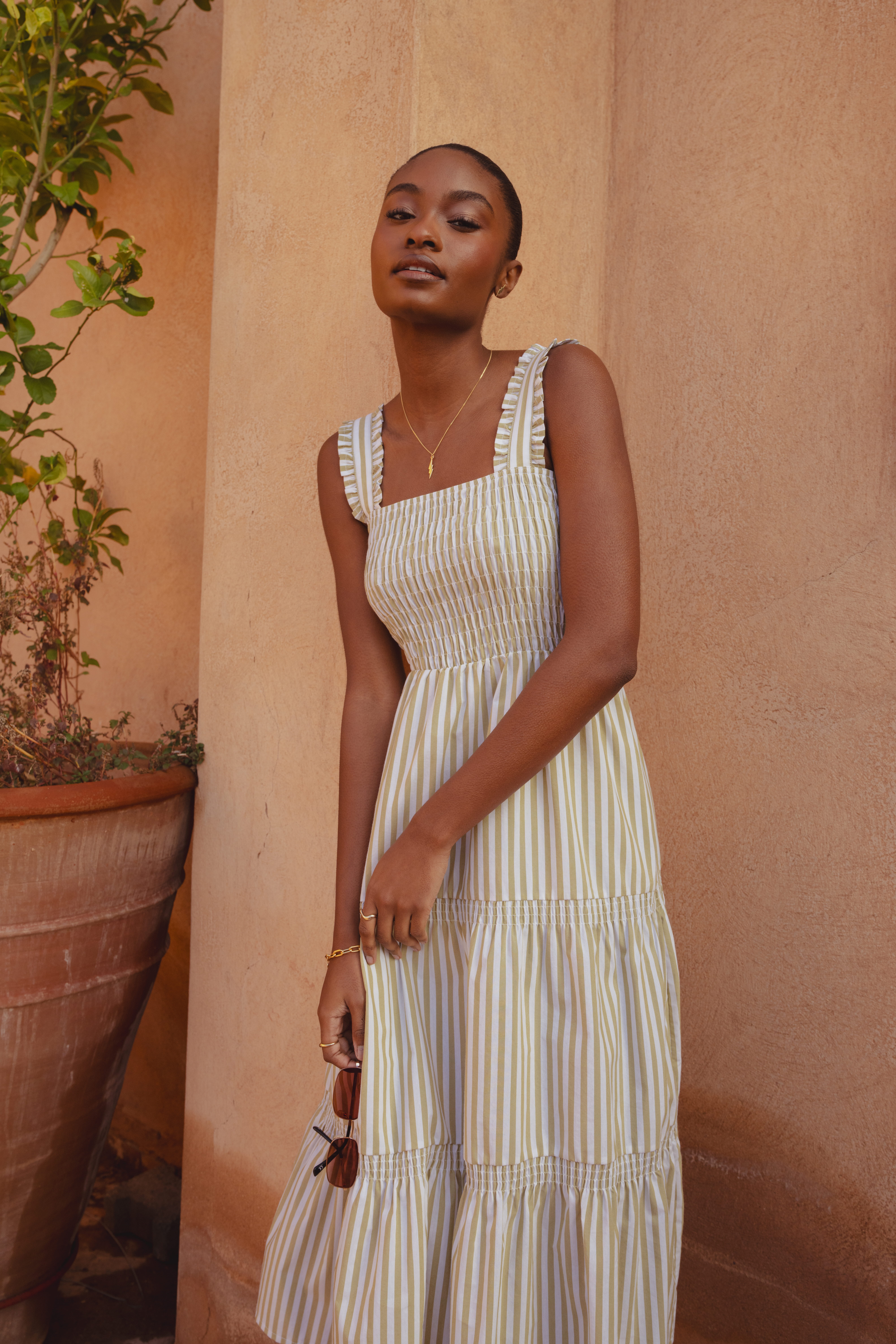 A model standing in front of a clay building wearing the Saint and Sofia Maya Shirring Dress in Green Stripe. This sleeveless dress features wide, ruffled straps with an elasticated bodice, breezy midi skirt with a tiered skirt for added fullness.