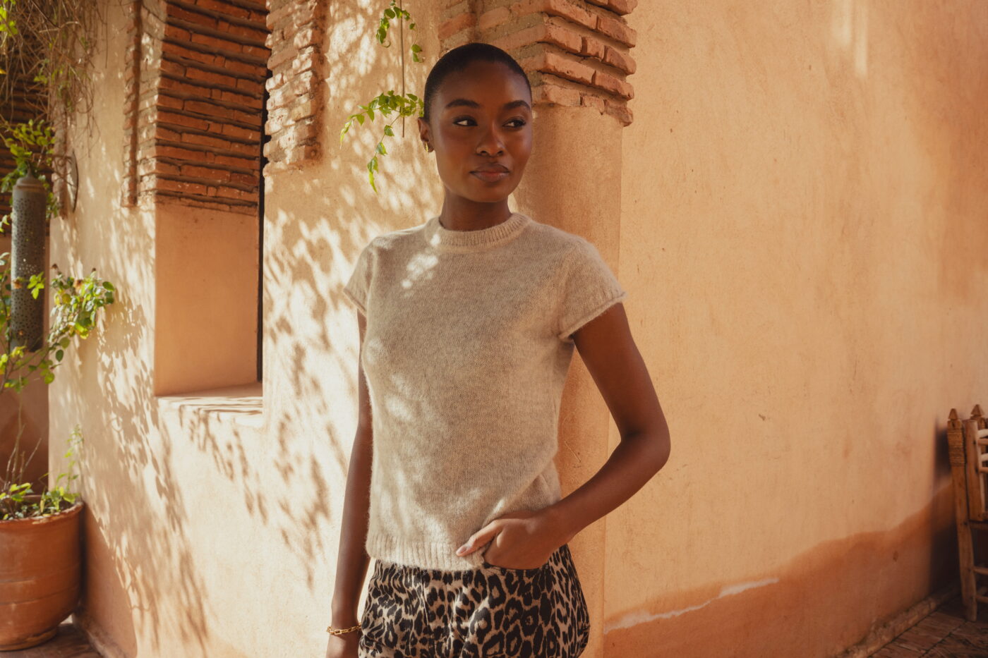 A model standing at the corner of a clay building looking into the distance. She wears the Saint and Sofia Ariella Baby Tee in Beige with the Leopard Print barrel leg jeans.