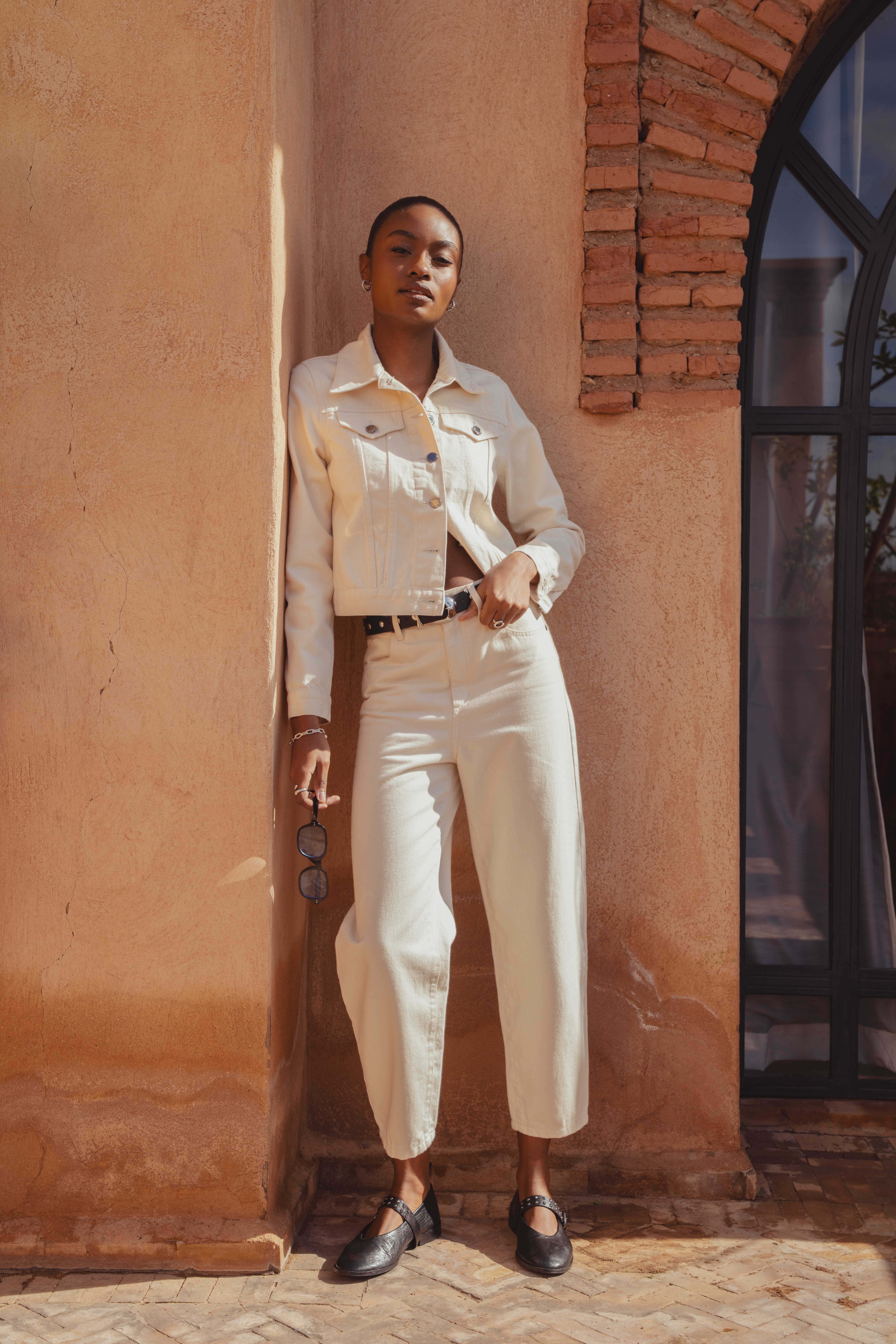 A model leaning against the side of a clay building wearing the Saint and Sofia Bowie Denim Jacket in Cream with the matching white Barrel Leg jeans. The look is styled with a black leather belt and black ballerina flats.