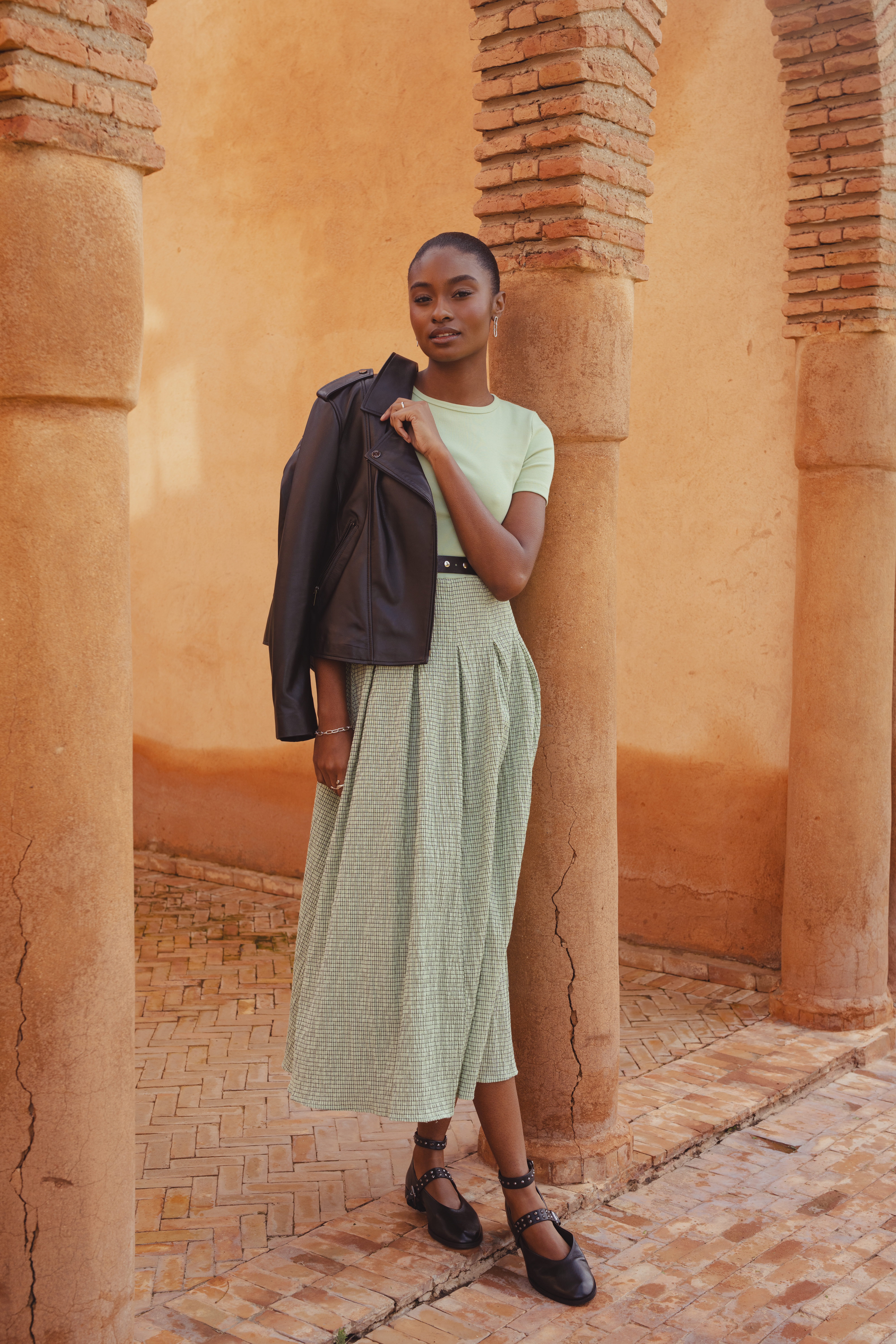 A model standing under a brick archway wearing the Saint and Sofia Renee Contrast Short Sleeve Dress in Mint Gingham. A co-ord effect midi dress with a short sleeve jersey bodice and gingham printed skirt. She styles it with the leather Luther Jacket, a leather and metal studded belt, and the leather Vega ballerinas.