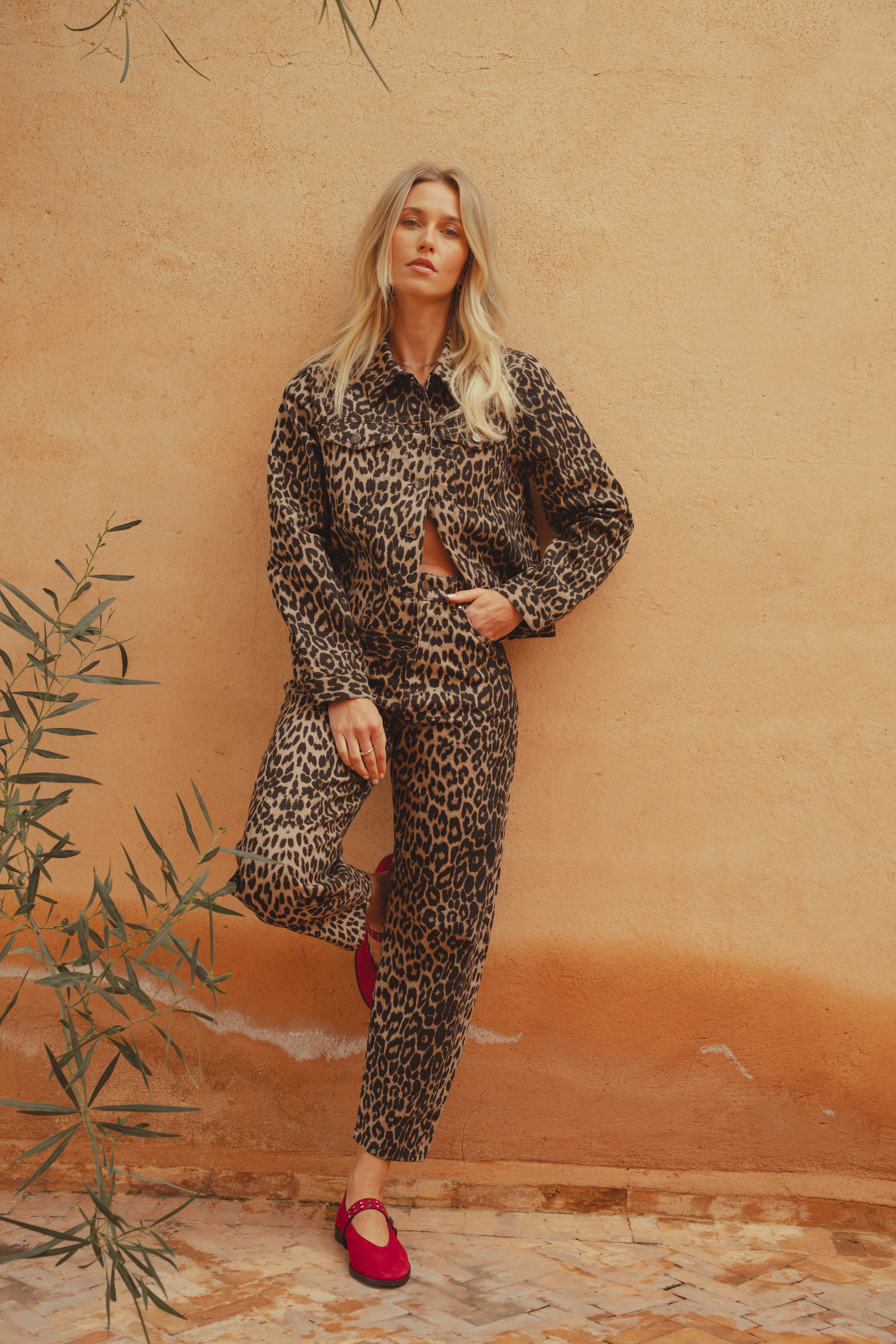 A model leaning against a clay wall with one leg bent at the knee and resting on the wall behind her. She wears the Saint and Sofia Bowie denim jacket in leopard print with the matching leopard print Barrel Leg Jeans. She styles the jacket open and pairs the look with the red Vega ballerina flats in suede.