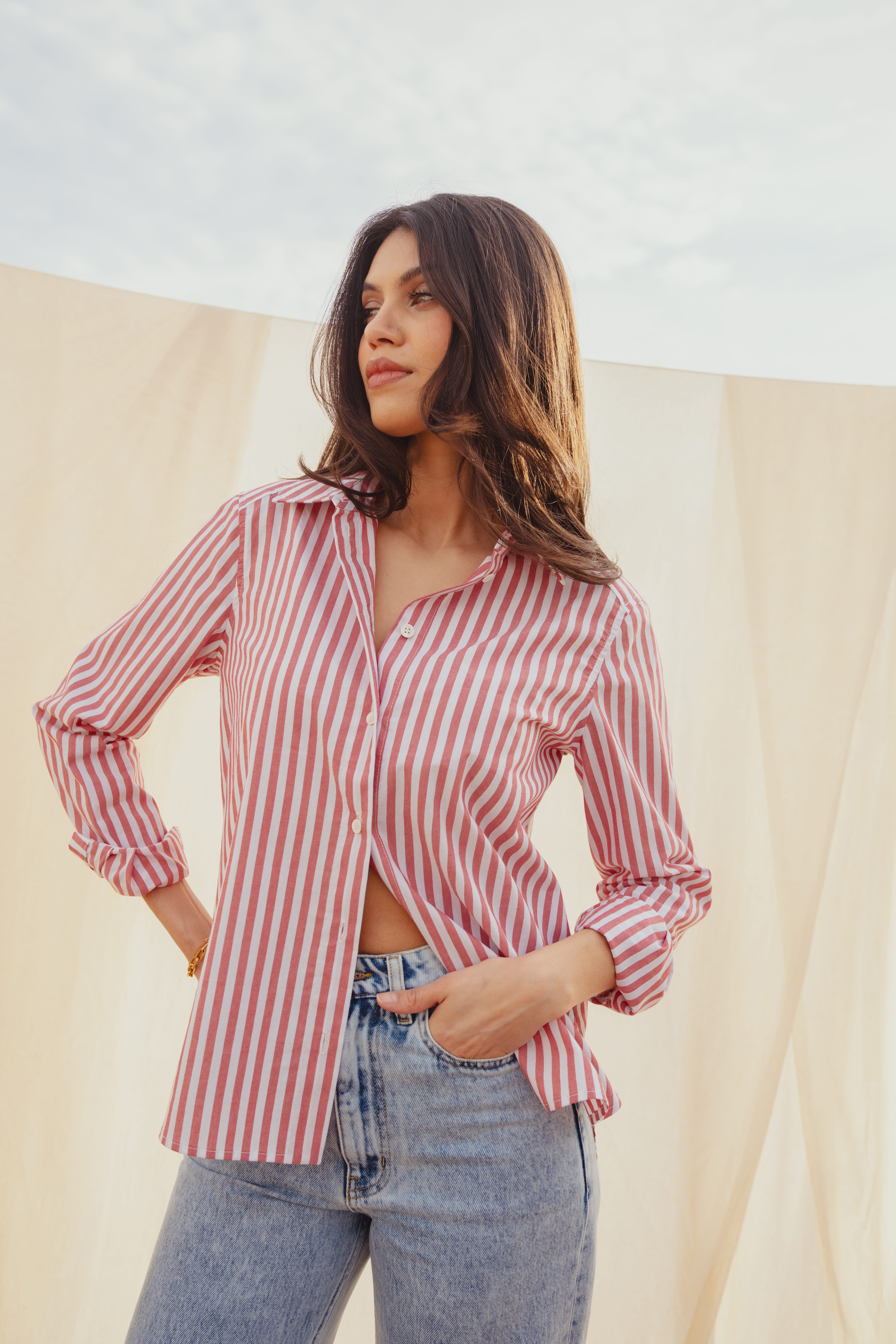 A model standing in front of a cream sheet that hangs over a clothes line. The blue sky is above her head. She wears a the red and white striped Anneleise shirt from Saint and Sofia and the Barrel Leg Jeans in Mid Blue from Saint and Sofia. She stands with one hand on her hip and the other tucked into her front pocket at she looks to the left.