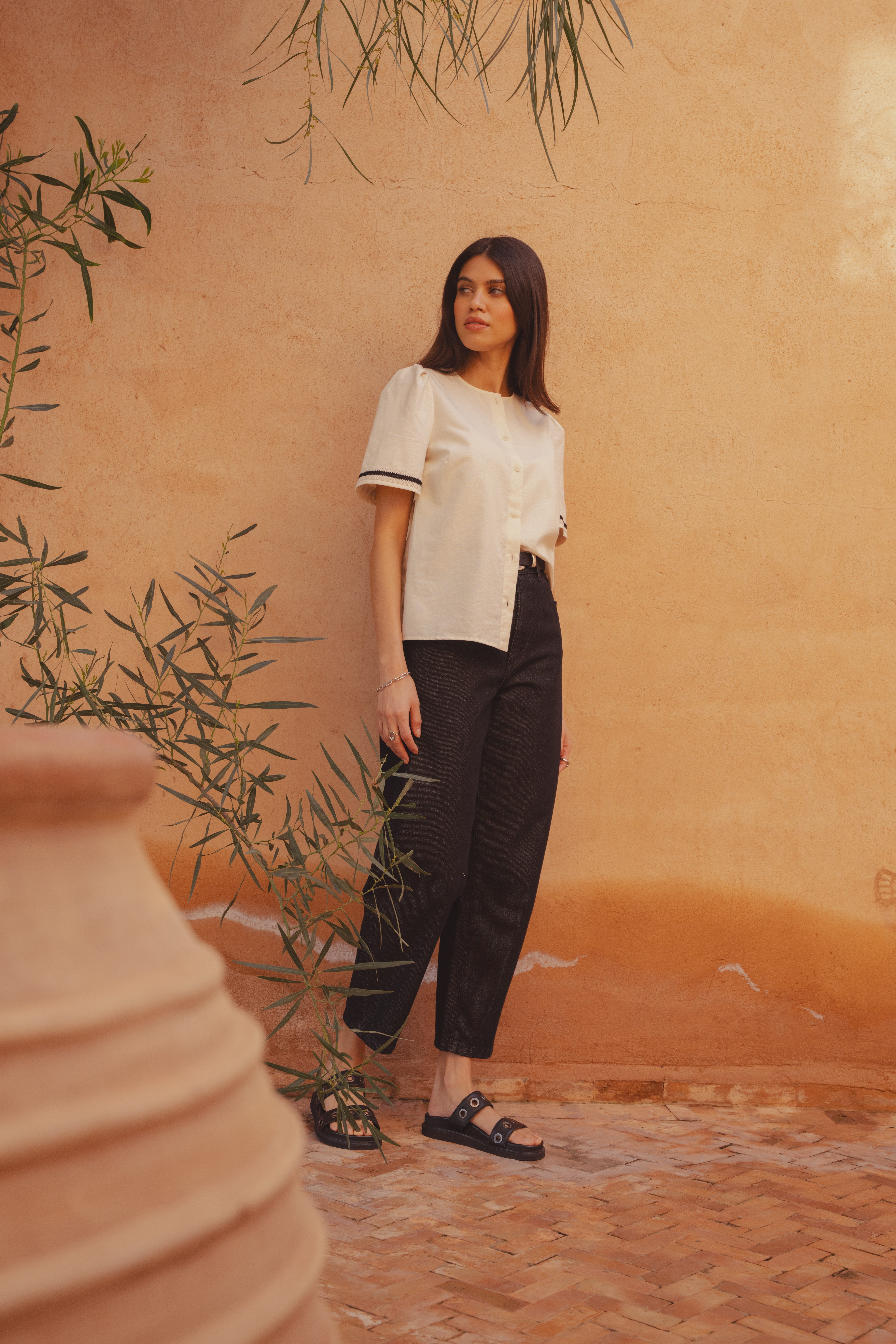 A model looking into the distance left of the camera, standing in front of a clay wall wearing the Saint and Sofia Tabitha blouse, a short sleeve button up style with one side of the hem tucked into a pair of the Barrel Leg Jeans in black. She styles the look with leather sliders.
