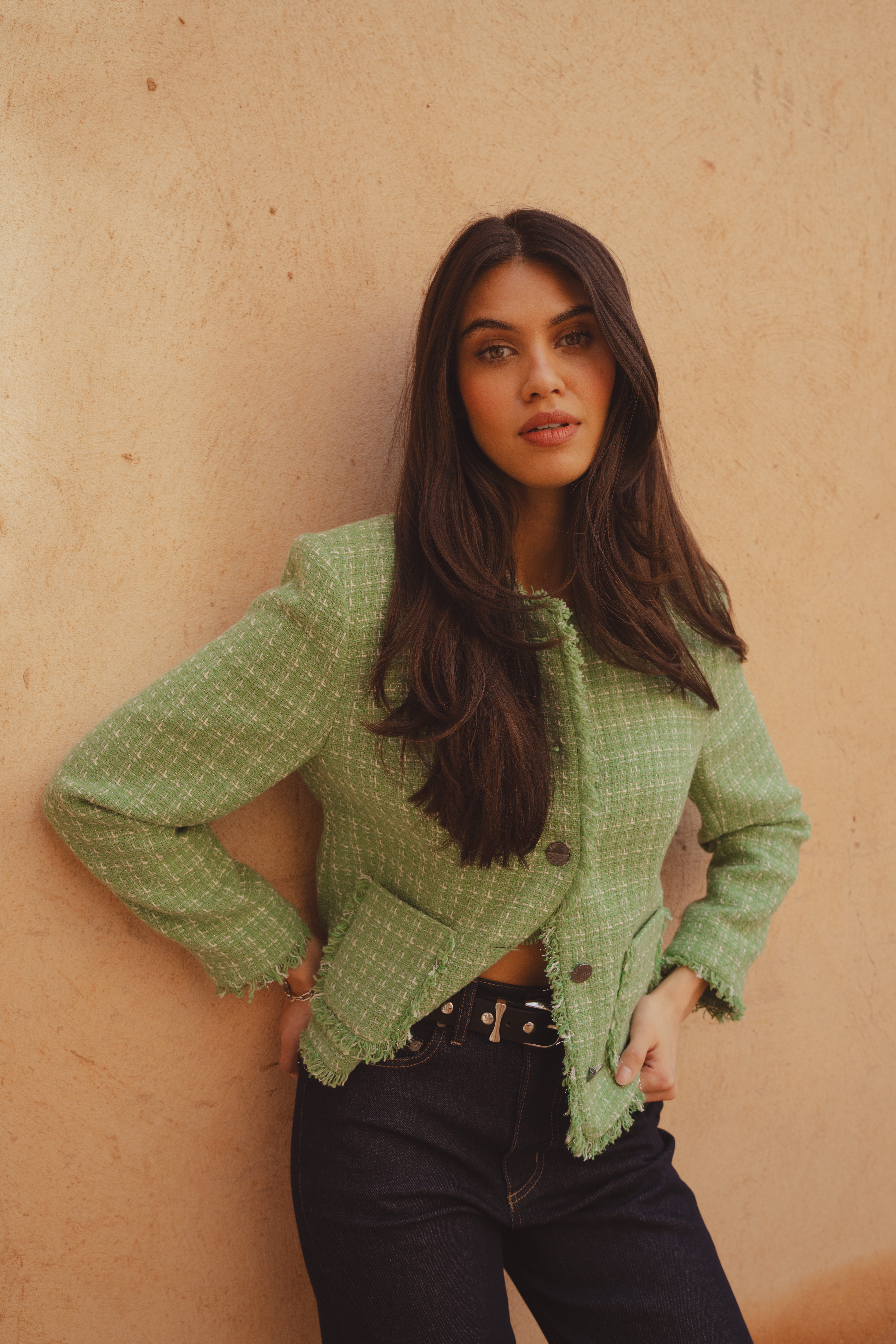 A model leaning against a clay building wearing the Saint and Sofia Ashby Jacket in Mint. A cropped boucle jacket with a frayed trim, front patch pockets, and gunmetal buttons. She wears it with a pair of Indigo barrel leg jeans and a black leather belt.