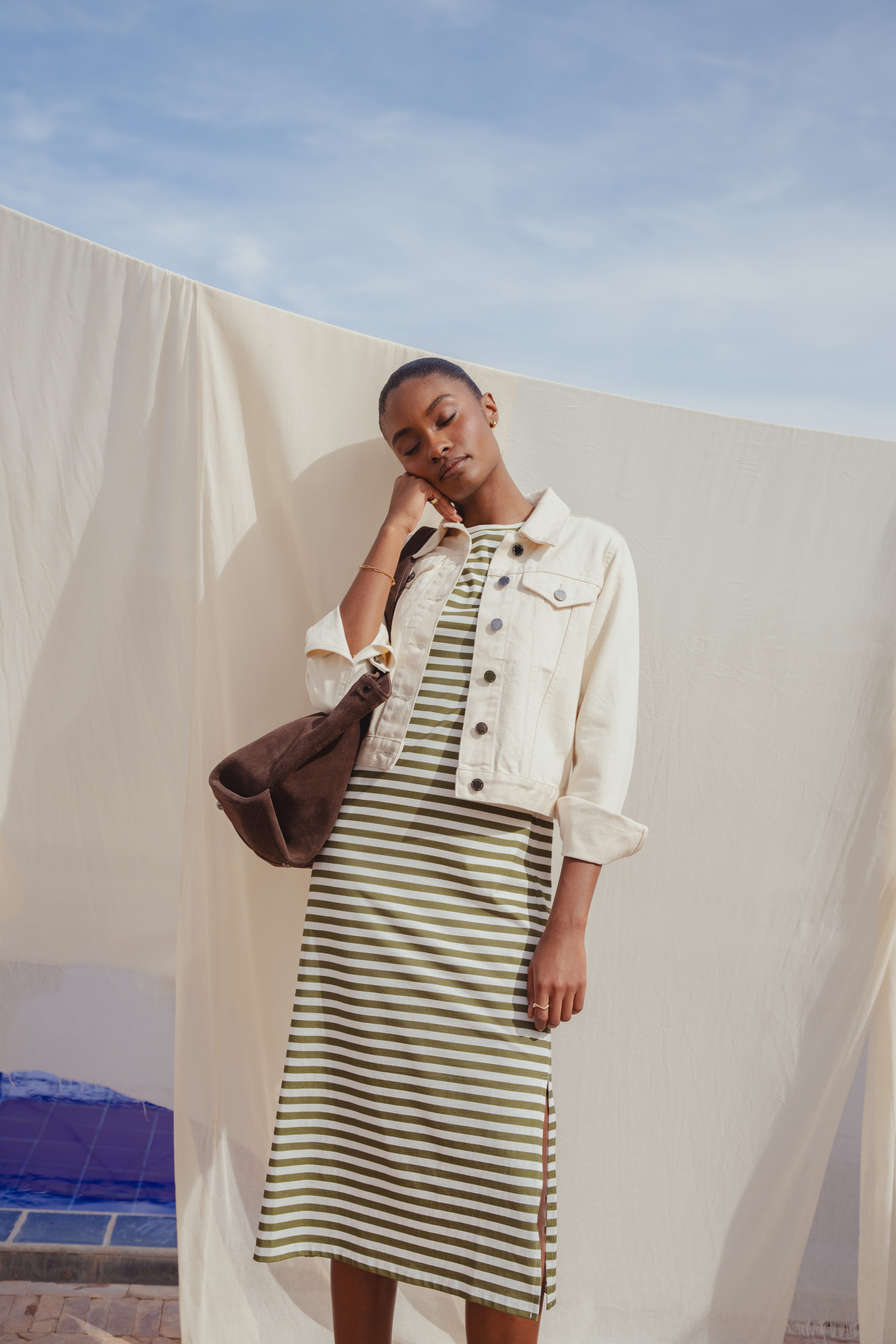 A model standing in front of a white fitted sheet that hangs outside under the sun. She wears the Saint and Sofia Easy T Shirt Dress, a crew neck style with green and white stripes. Over the top, she pairs it with a white denim jacket and finishes the look with a khaki suede shoulder bag.