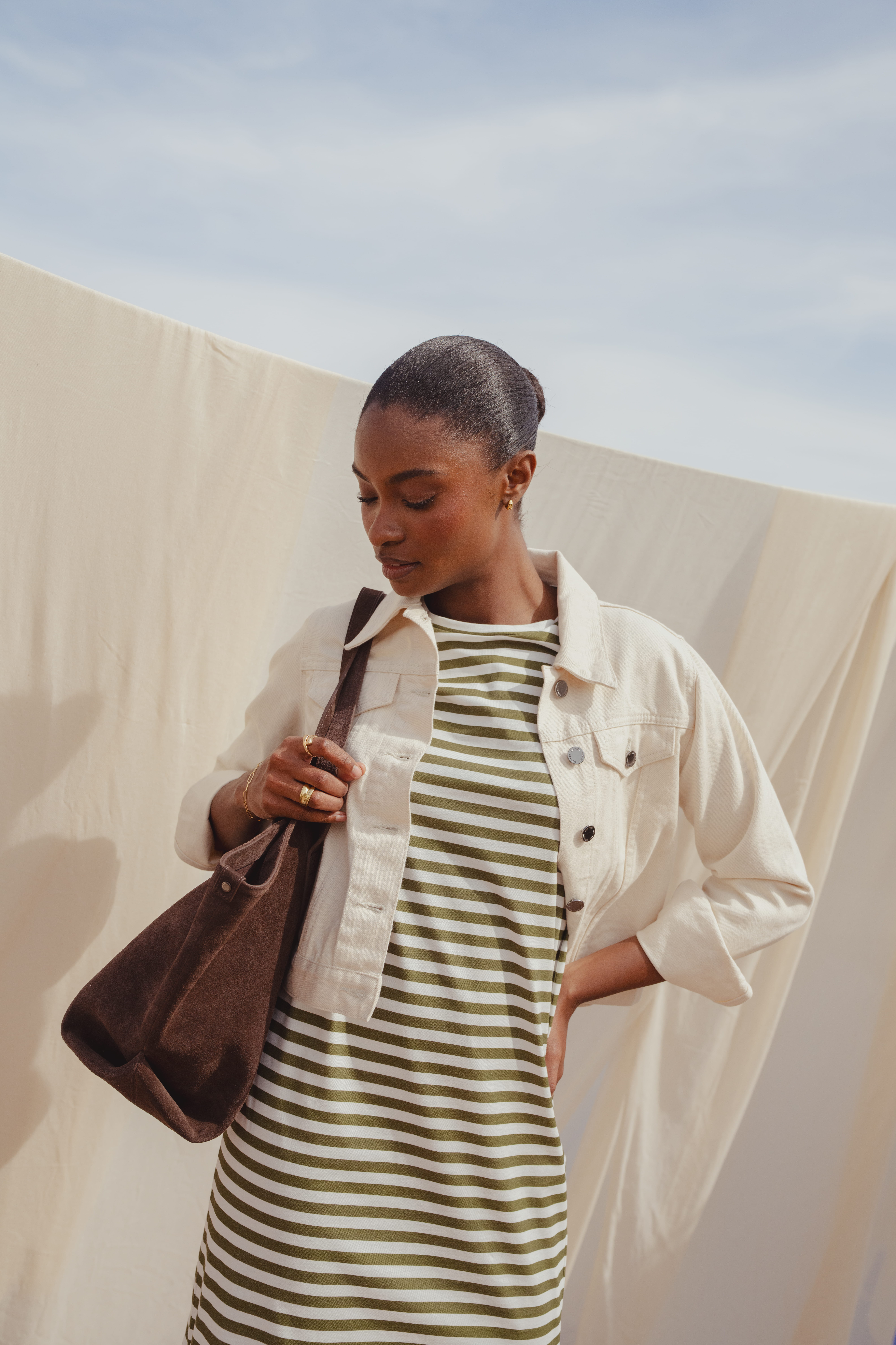 A model standing in front of a white fitted sheet that hangs outside under the sun. She wears the Saint and Sofia Easy T Shirt Dress, a crew neck style with green and white stripes. Over the top, she pairs it with a white denim jacket and finishes the look with a khaki suede shoulder bag.