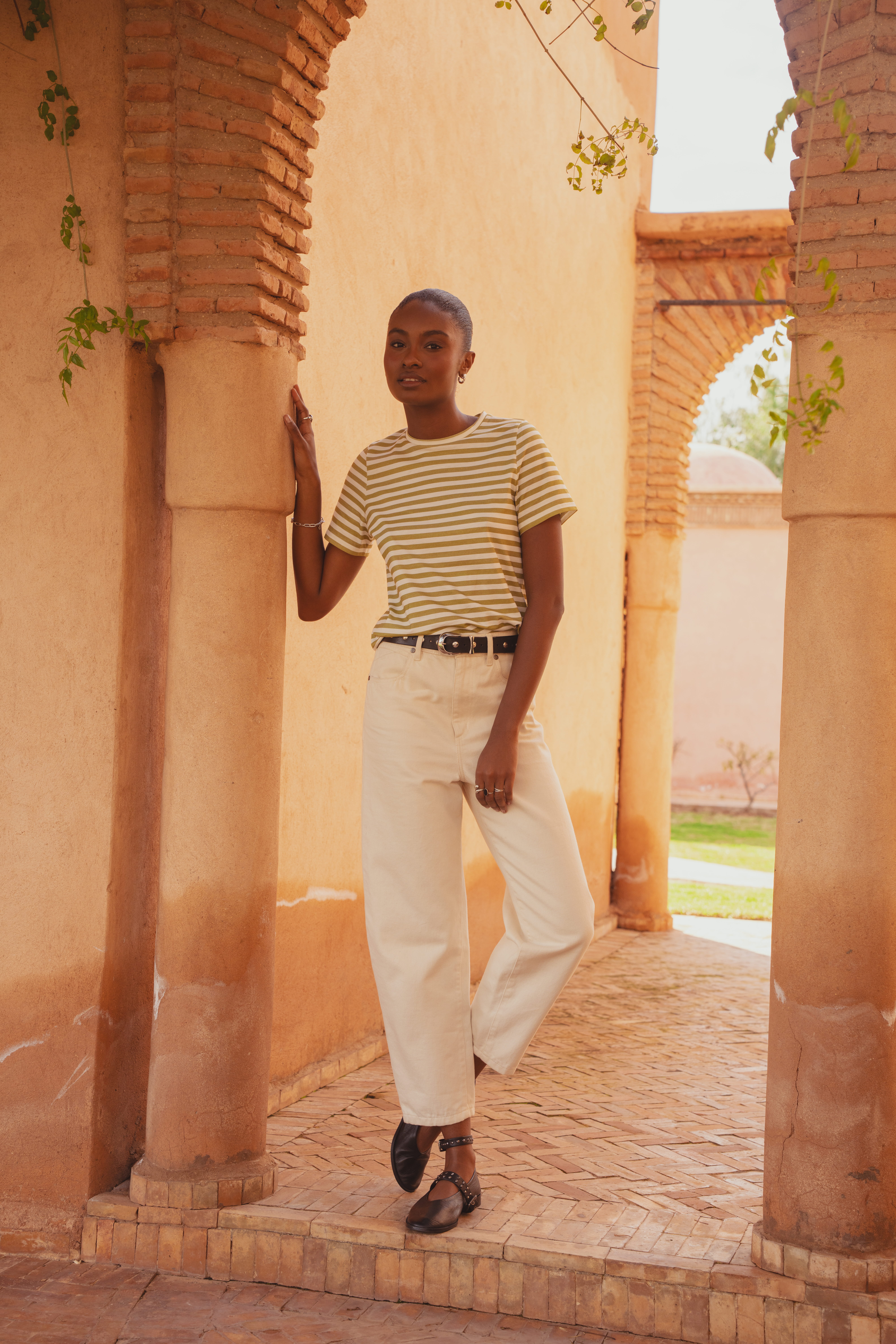 A model standing under the archway of a clay building wearing the Saint and Sofia Chelsea Crew Neck Tee with green and white stripes tucked into a pair of the Barrel Leg Jeans. She styles it with a leather belt and leather ballerina flats.