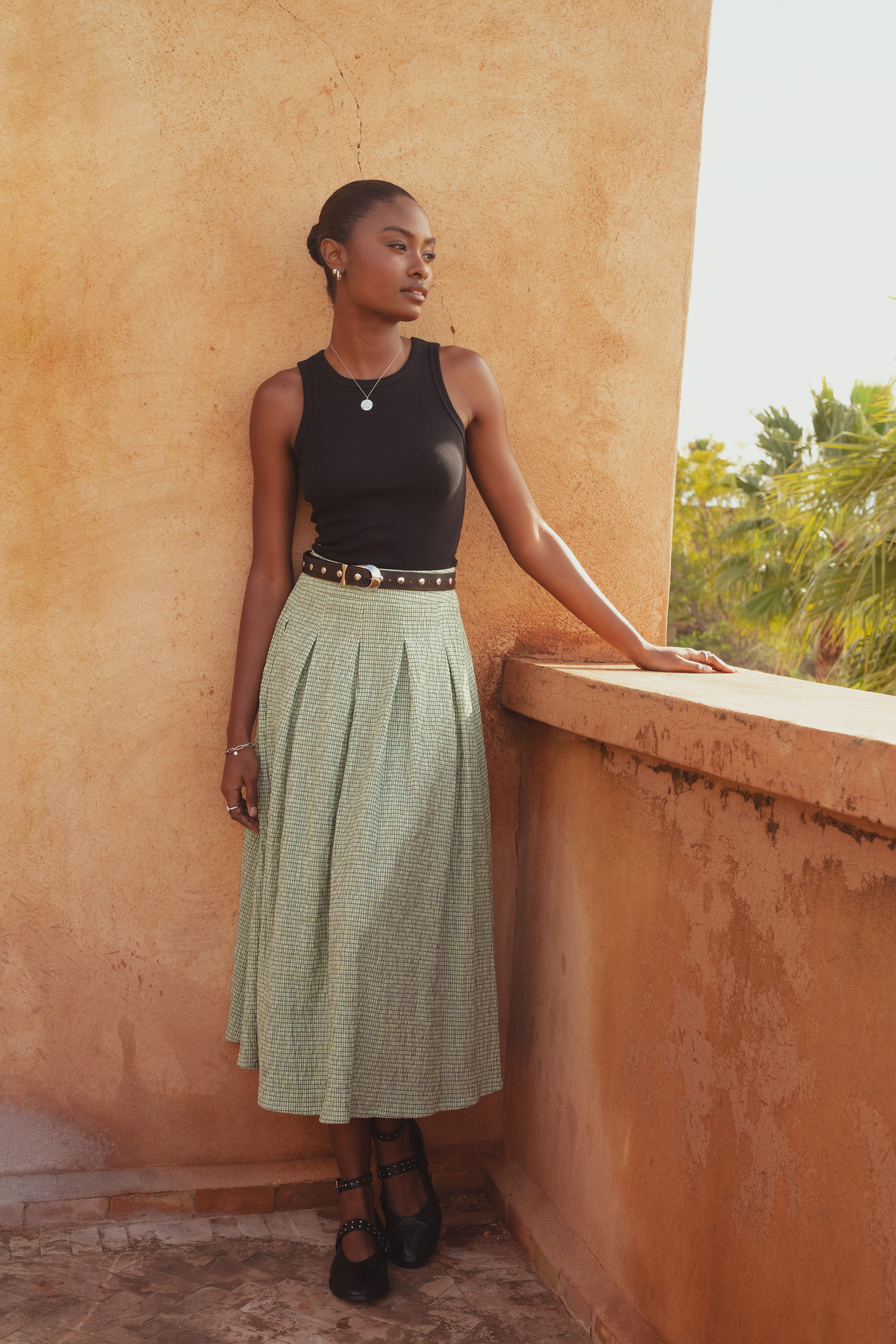 A model standing on a balcony with her hand resting against the concrete railing. She looks out to the distance and wears the Saint and Sofia Eliza Pleated Skirt in Mint Gingham, a midi skirt design with box pleats and a mid-rise waistband. She styles it with a black racer vest tee and black leather ballerina flats.