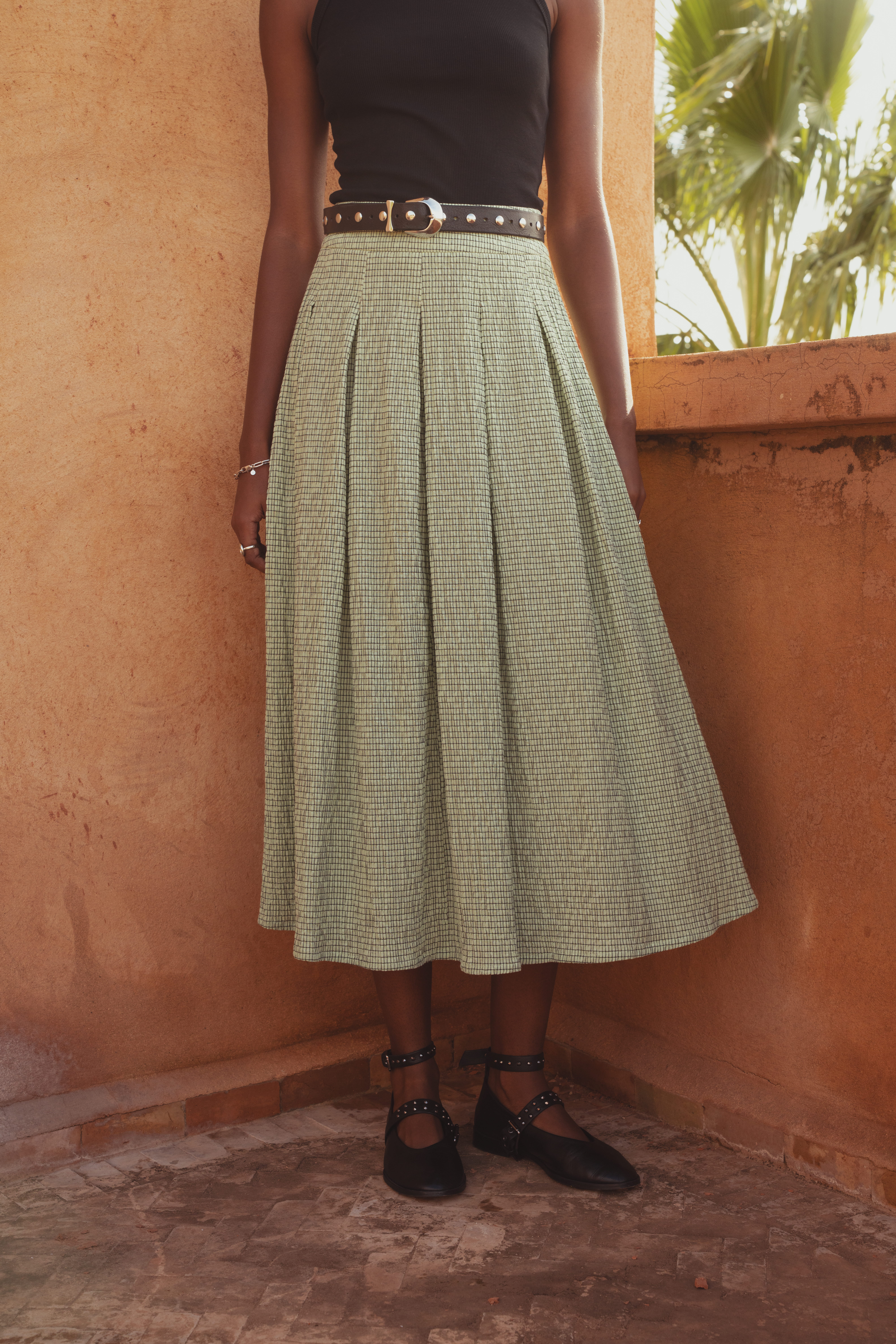 An up-close shot of the Saint and Sofia Eliza Pleated Skirt in Mint Gingham. A midi skirt design with box pleats. It's styled here with a studded leather belt and a black tank top. Then paired with leather ballerina flats.