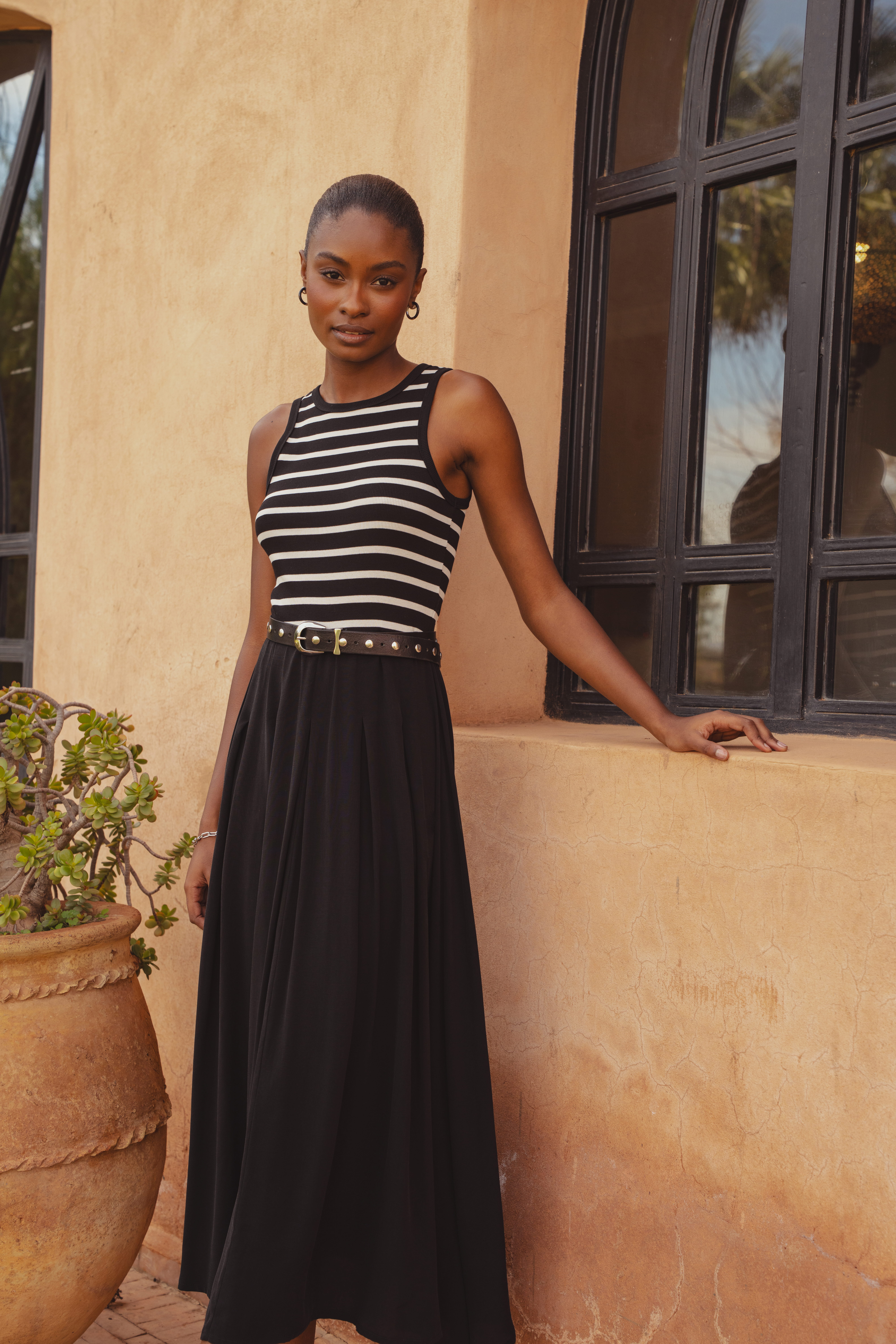 A model standing in front of a clay building with her arm resting on the window frame behind her. She wears the Saint and Sofia Renee Contrast Dress, a sleeveless style with a black and white striped bodice and black flowy skirt.