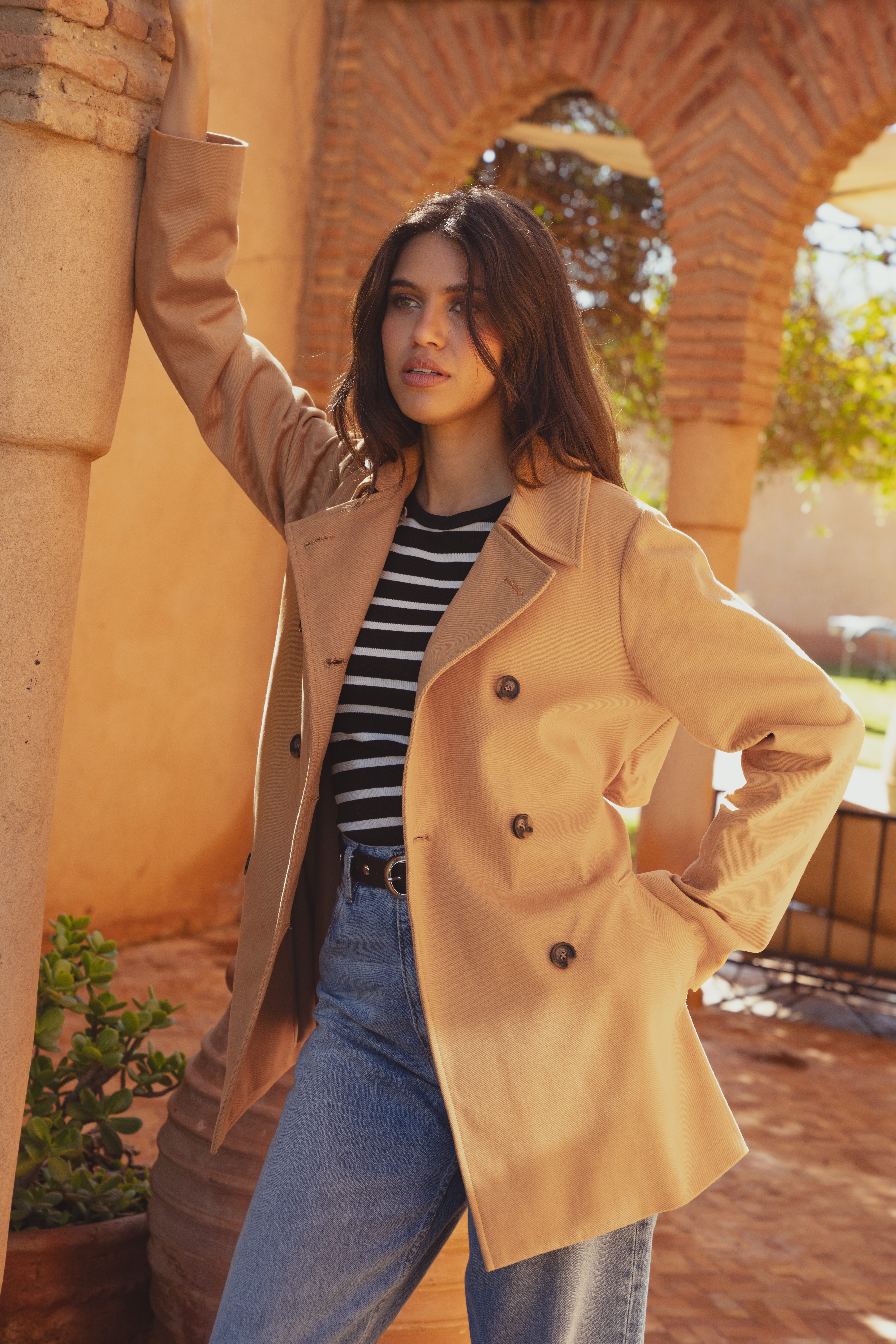A model leaning against a pillar wearing the Saint and Sofia cropped trench coat. The Marion trench coat is a double breasted style in a camel colour. She wears it here with a striped black and white long sleeved top and a pair of light blue jeans.