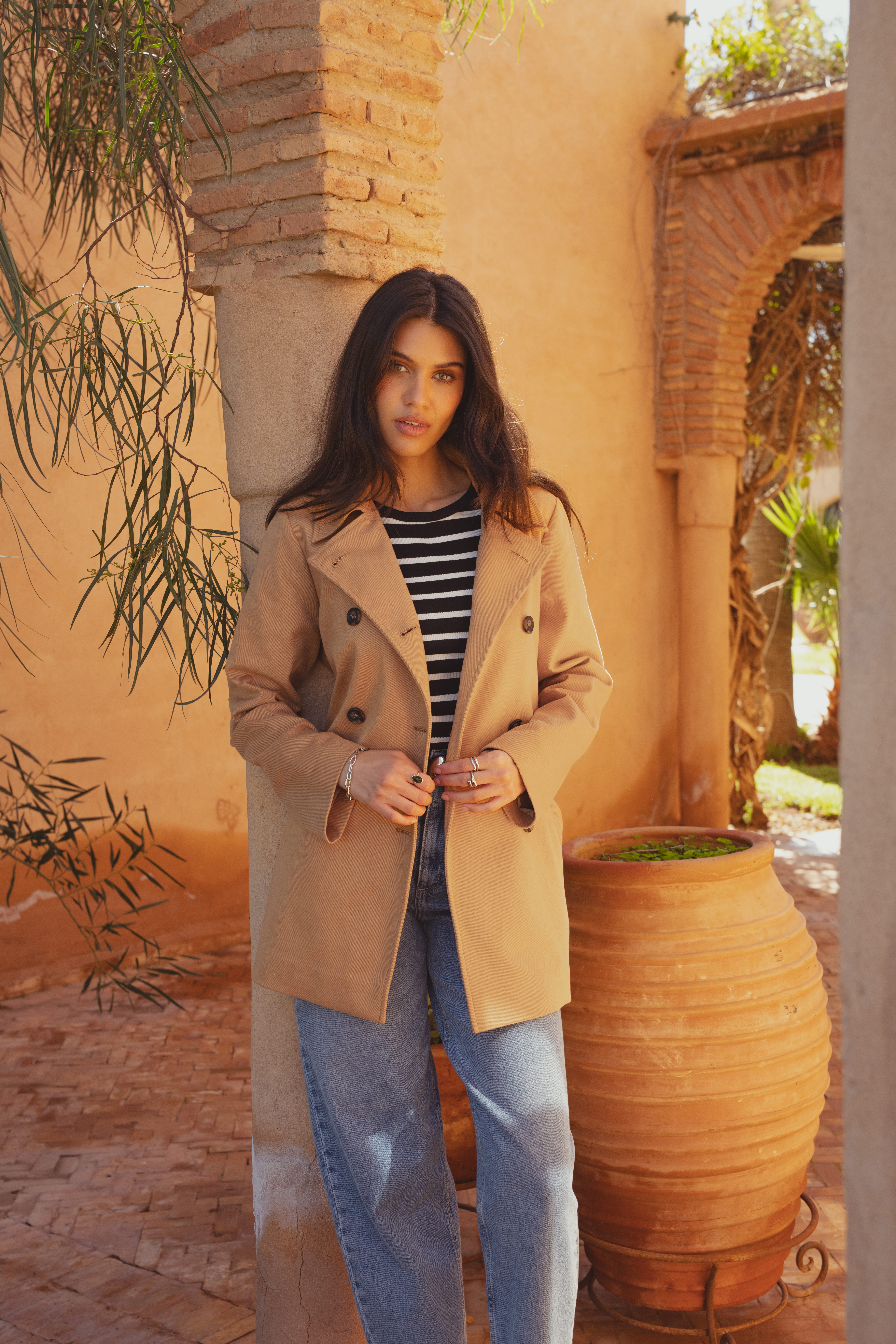 A model leaning against a pillar wearing the Saint and Sofia cropped trench coat. The Marion trench coat is a double breasted style in a camel colour. She wears it here with a striped black and white long sleeved top and a pair of light blue jeans.