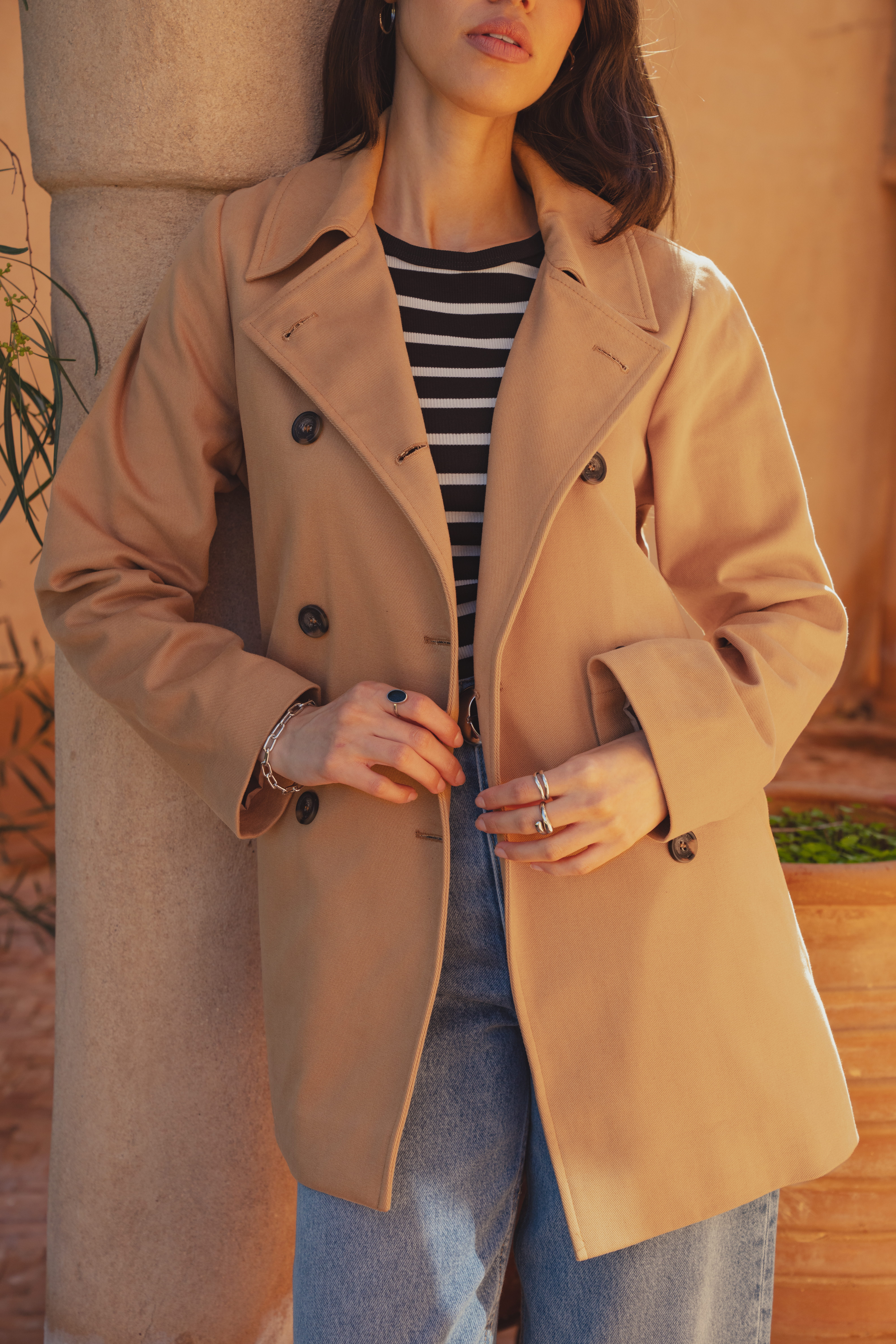 A close up image of the front of the Marion Short Trench Coat by Saint and Sofia. This cropped style is a classic camel colour in a traditional double breasted cut with button fastening and a collared neckline. The model wears it here with a black and white striped top and mid blue jeans.