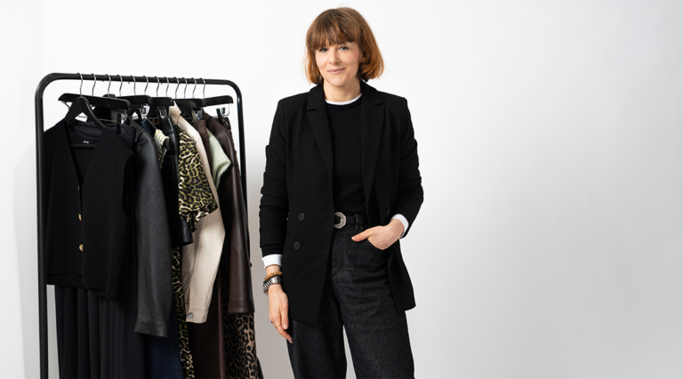 An image of the Saint and Sofia Head of Editorial, Alexandra Fullerton, wearing a black blazer with black trousers and a black crew neck tee. She stands in front of a white background and beside a black rail that displays clothes from Saint and Sofia's latest collection.