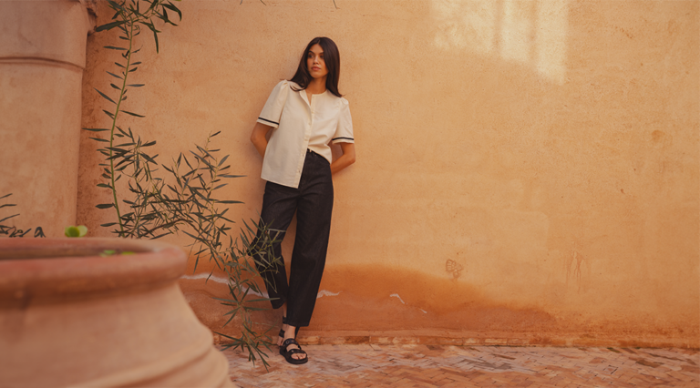 An image of a model leaning against a clay wall wearing the Saint and Sofia Tabitha Blouse in Cream and the Barrel Leg Jeans in Black with the leather Madison Eyelet slides. She looks out to the left as the photo is taken.
