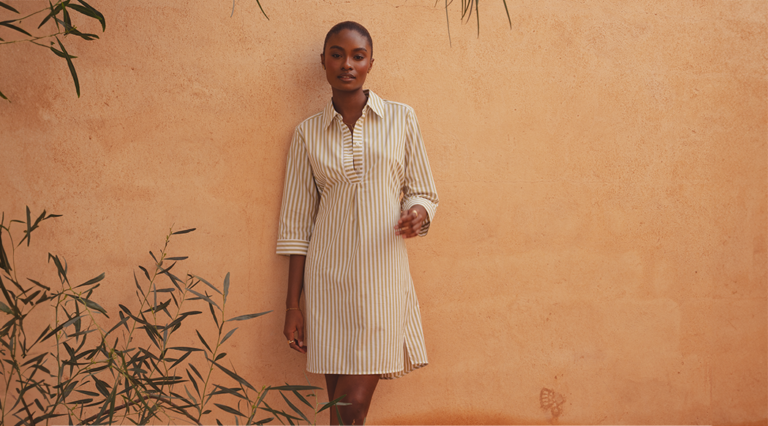 A model standing against a clay wall wearing a green and white shirt dress from Saint and Sofia. It's made with an open collared neckline, three quarter length sleeves, and a mini skirt length