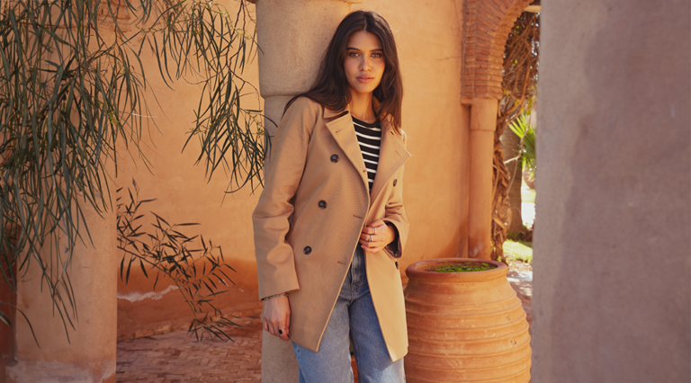 A model leaning against a pillar wearing a cropped trench coat by saint and sofia. This is a double breasted cropped trench coat style in a camel colour. She wears it here with a pair of light blue denim jeans.