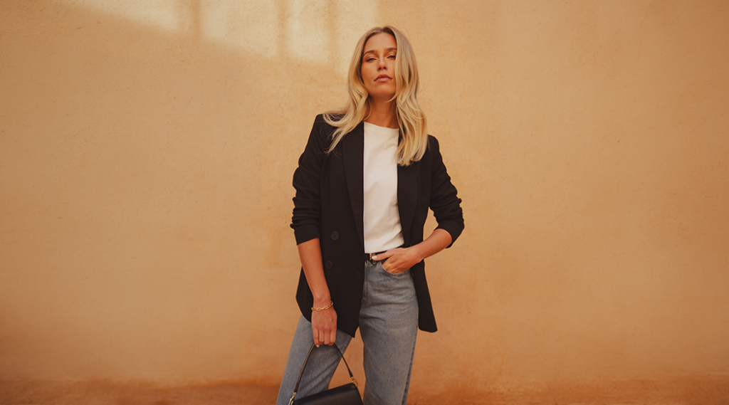A model standing in front of a clay wall wearing a black blazer from saint and sofia with a white crew neck tee and a pair of mid-blue barrel leg jeans. In her hand, she holds a black leather bag