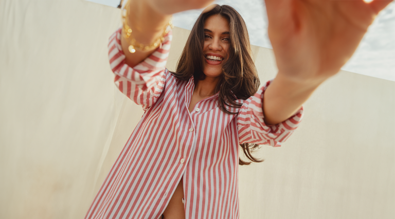 A model standing in front a cream sheet that hangs over a clothes line. She wears a red and white Saint and Sofia striped shirt, unbuttoned at the hem to leave a glimpse of her stomach on show. She stands with her hands outstretched toward the camera as she smiles down the lens.