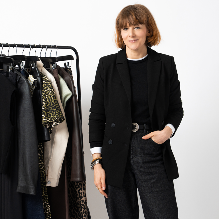 An image of the Saint and Sofia Head of Editorial, Alexandra Fullerton, wearing a black blazer with black trousers and a black crew neck tee. She stands in front of a white background and beside a black rail that displays clothes from Saint and Sofia's latest collection.