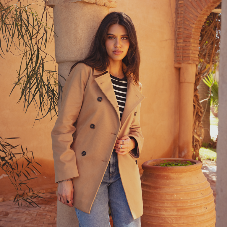 A model leaning against a pillar wearing a cropped trench coat by saint and sofia. This is a double breasted cropped trench coat style in a camel colour. She wears it here with a pair of light blue denim jeans.