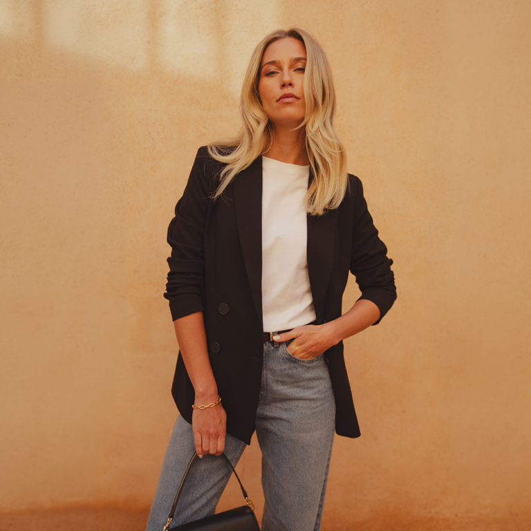 A model standing in front of a clay wall wearing a black blazer from saint and sofia with a white crew neck tee and a pair of mid-blue barrel leg jeans. In her hand, she holds a black leather bag