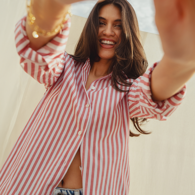 A model standing in front a cream sheet that hangs over a clothes line. She wears a red and white Saint and Sofia striped shirt, unbuttoned at the hem to leave a glimpse of her stomach on show. She stands with her hands outstretched toward the camera as she smiles down the lens.