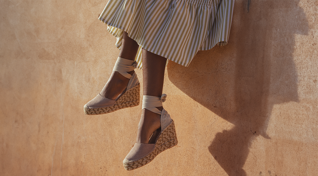 A model's legs. She sits on a wall and her legs dangle into shot. She wears a striped midi dress from Saint and Sofia and a pair of wedge heeled espadrilles the tie around the ankle.