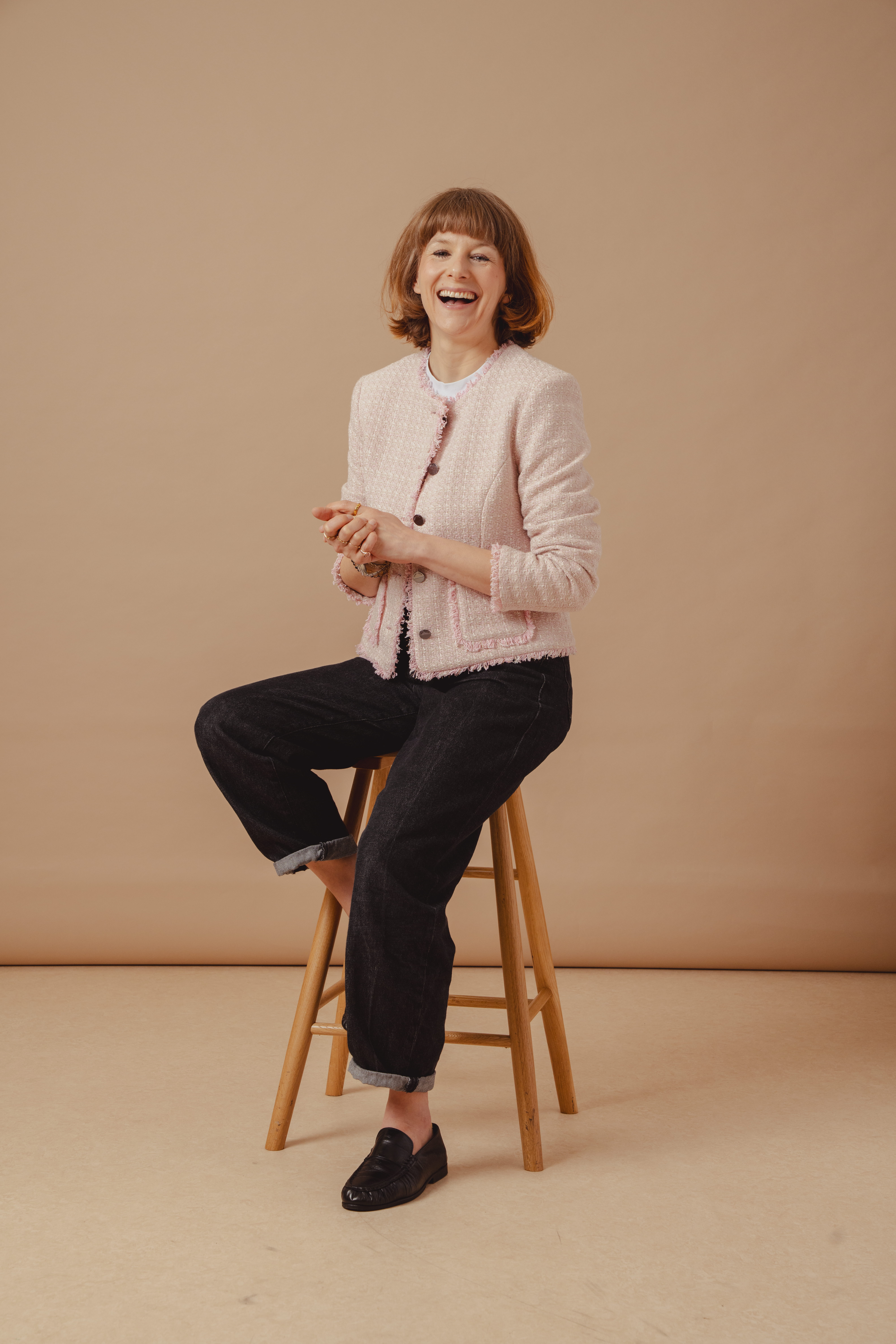 Saint and Sofia's Head of Editorial, Alexandra Fullerton, sat on a stool in front of a beige background. She wears the pink Rayner Jacket over a white tee with Indigo denim jeans and black trainers.