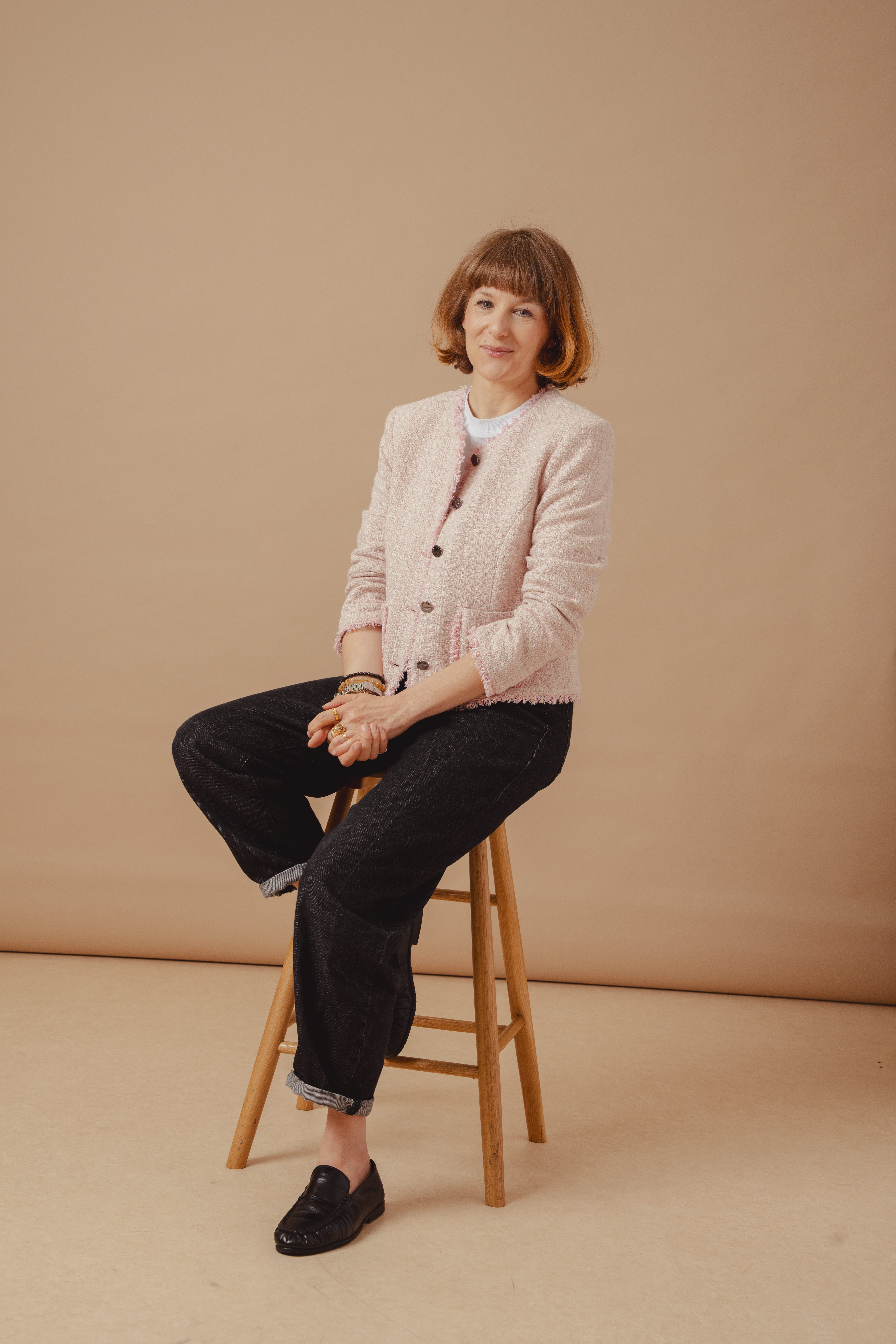 Saint and Sofia's Head of Editorial, Alexandra Fullerton, sat on a stool in front of a beige background. She wears the pink Rayner Jacket over a white tee with Indigo denim jeans and black trainers.