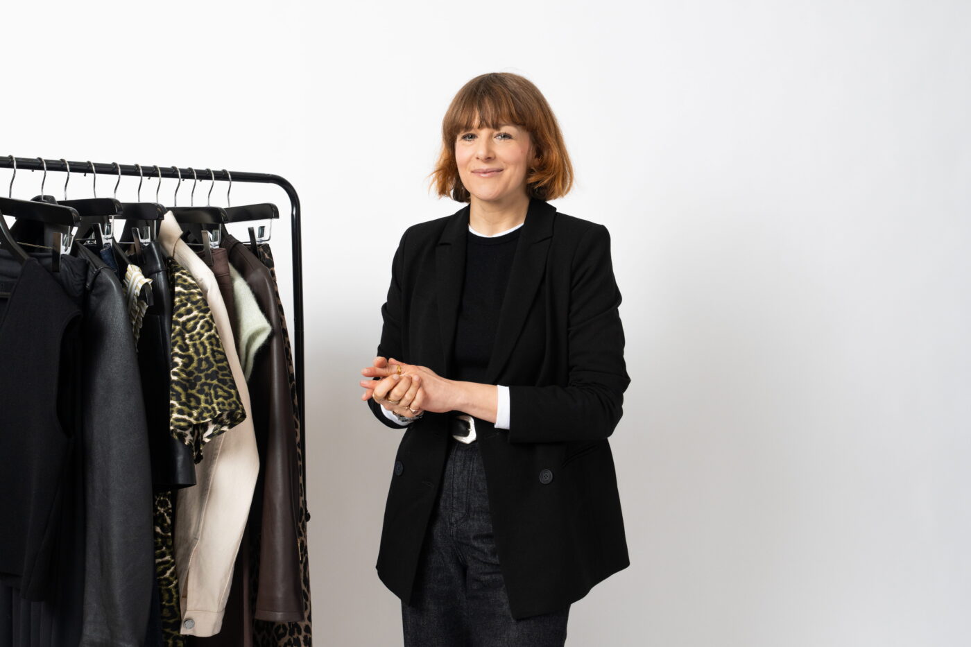 Alexandra Fullerton the Head of Editorial at Saint and Sofia stood with her hands clasped in front of her chest. She wears a black blazer over a black sweater and black jeans. She is standing beside a rail of Saint and Sofia clothes.