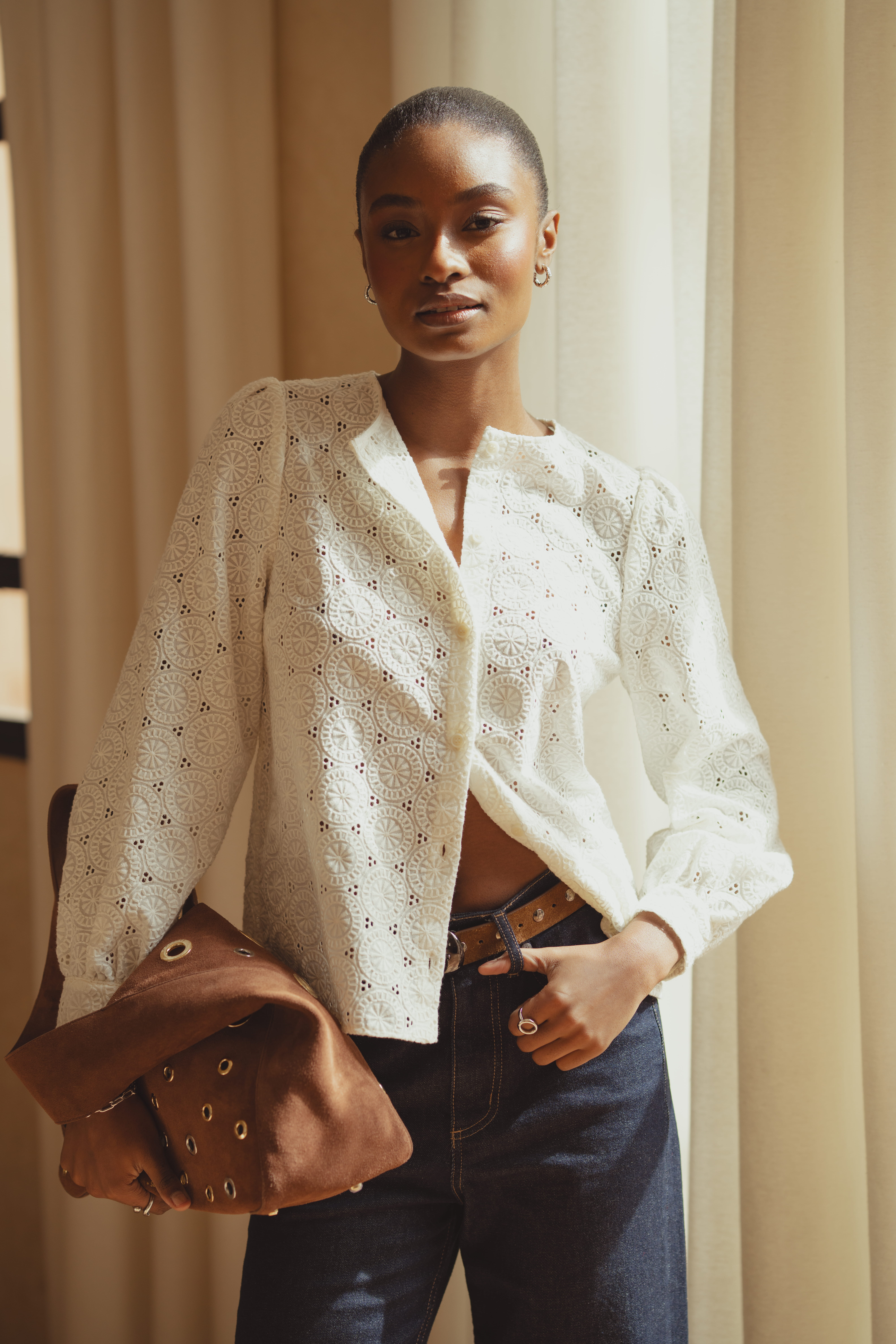 A model wearing the Tabitha blouse by Saint and Sofia a crew neck button-up style. She wears it undone at the hem with a pair of indigo blue wide leg jeans and a brown suede belt. In her hand, she holds the suede Allana Eyelet Bag.