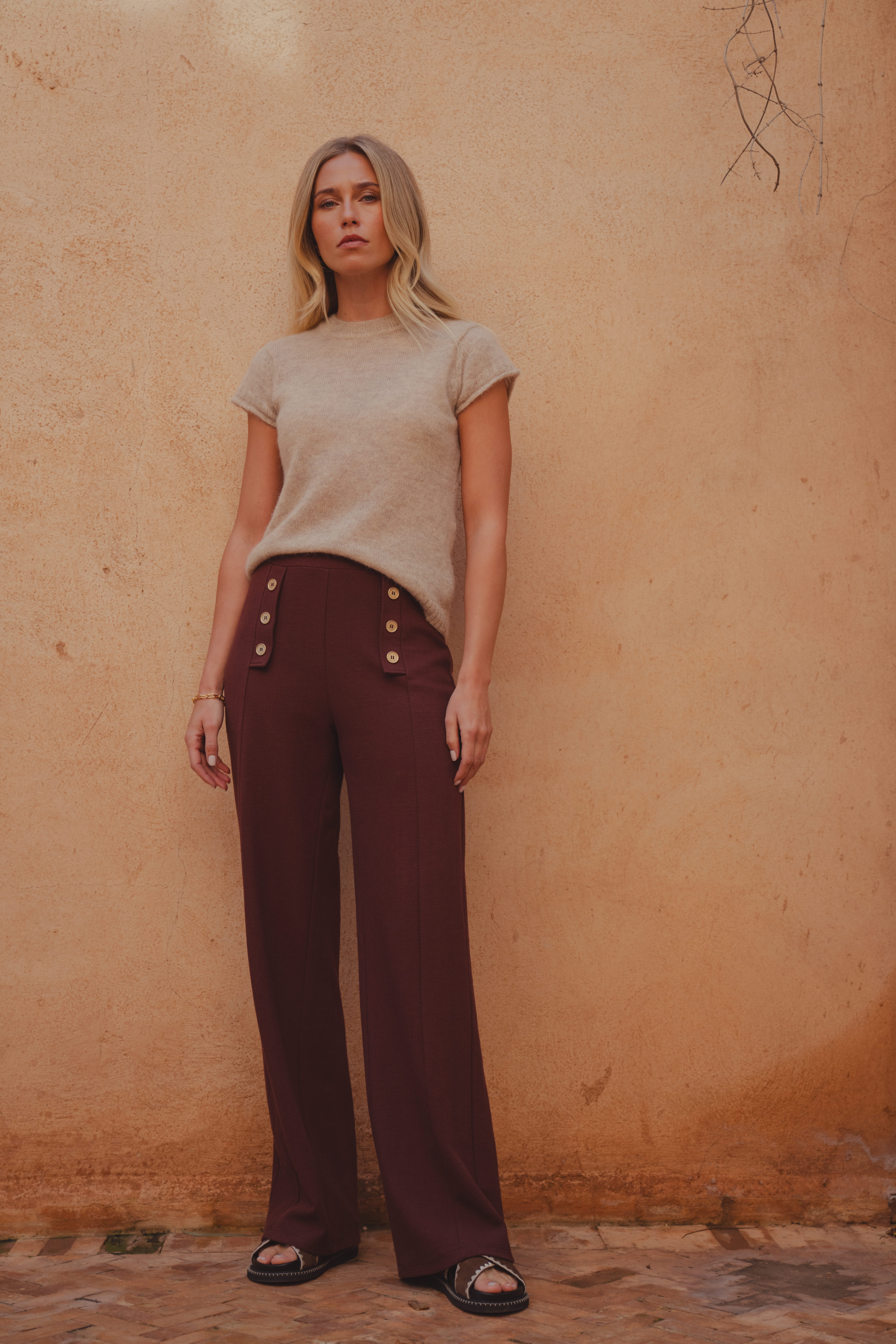 A model wearing a short sleeve beige knitted top with a crew neckline and a pair of brown wide leg trousers with gold button detail at the front. She styles it with a pair of brown slider sandals.