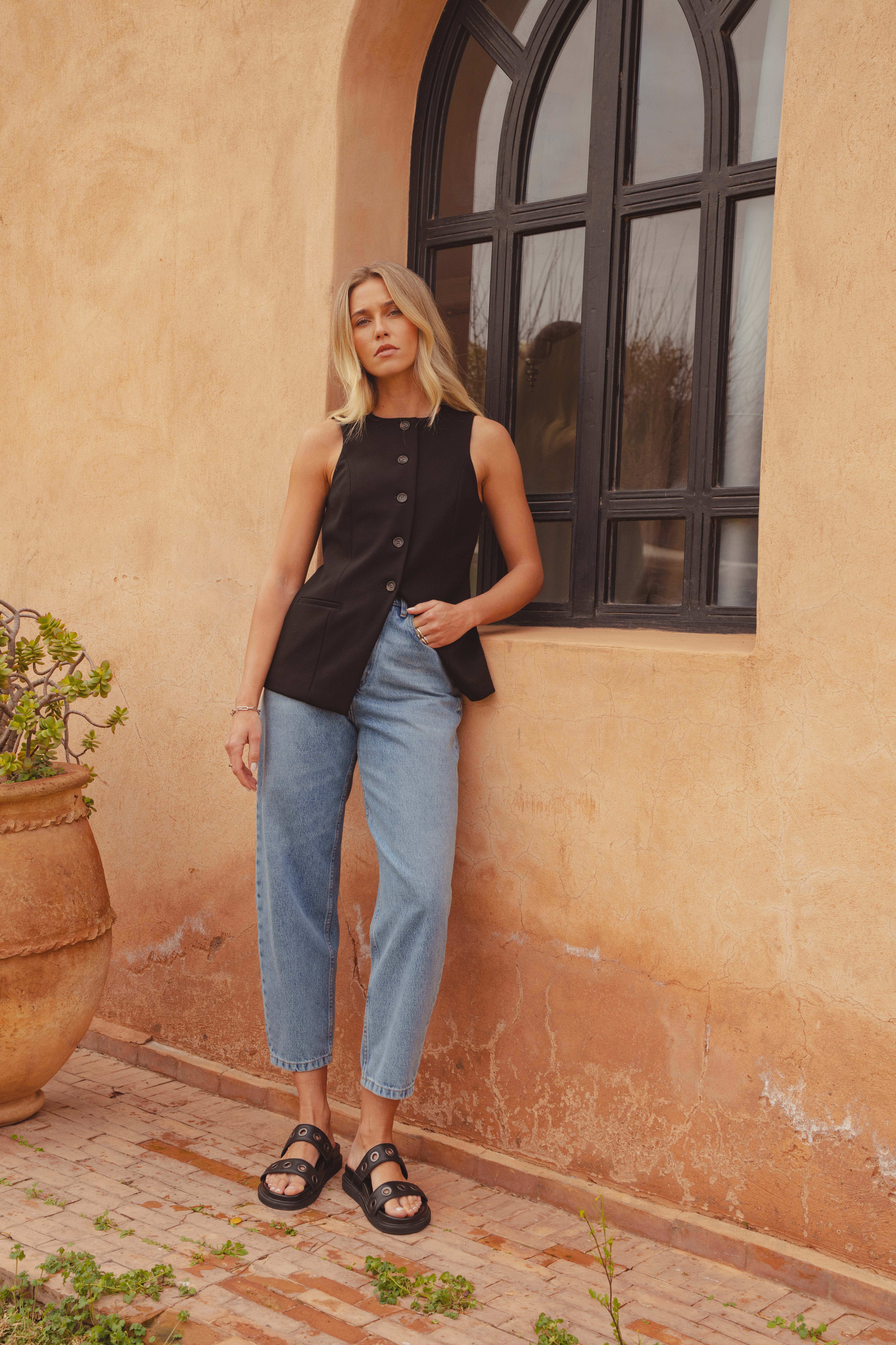 A model wearing the Argent waistcoat by saint and sofia in Black. A long line design with a crew neckline. It's worn here with mid blue barrel leg jeans and elather sliders.