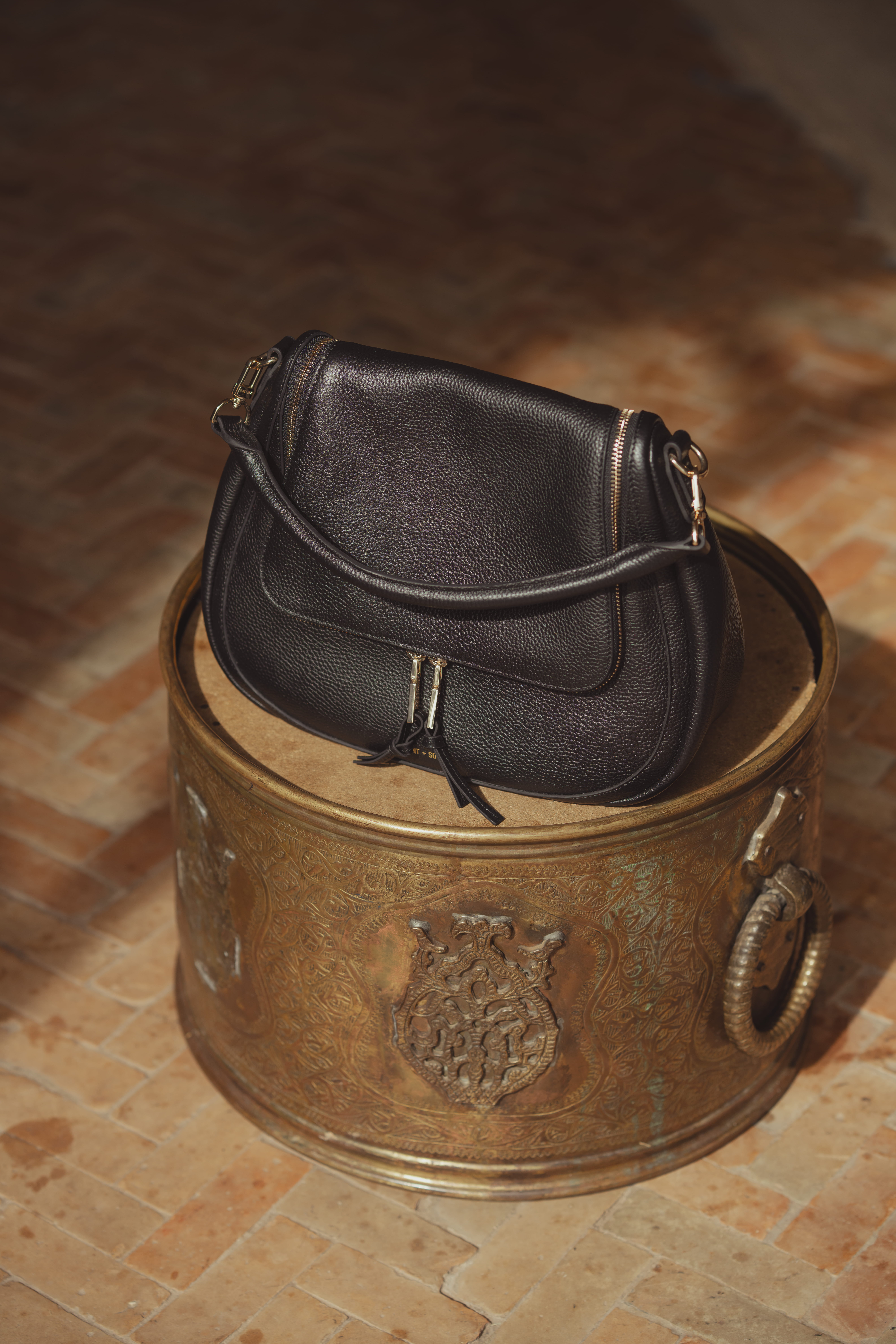 The Marlowe messenger bag by Saint and Sofia. A slouchy leather messenger style with a curved shape, top handle, and removable cross-body strap. It's photographed here on a copper metal stool.