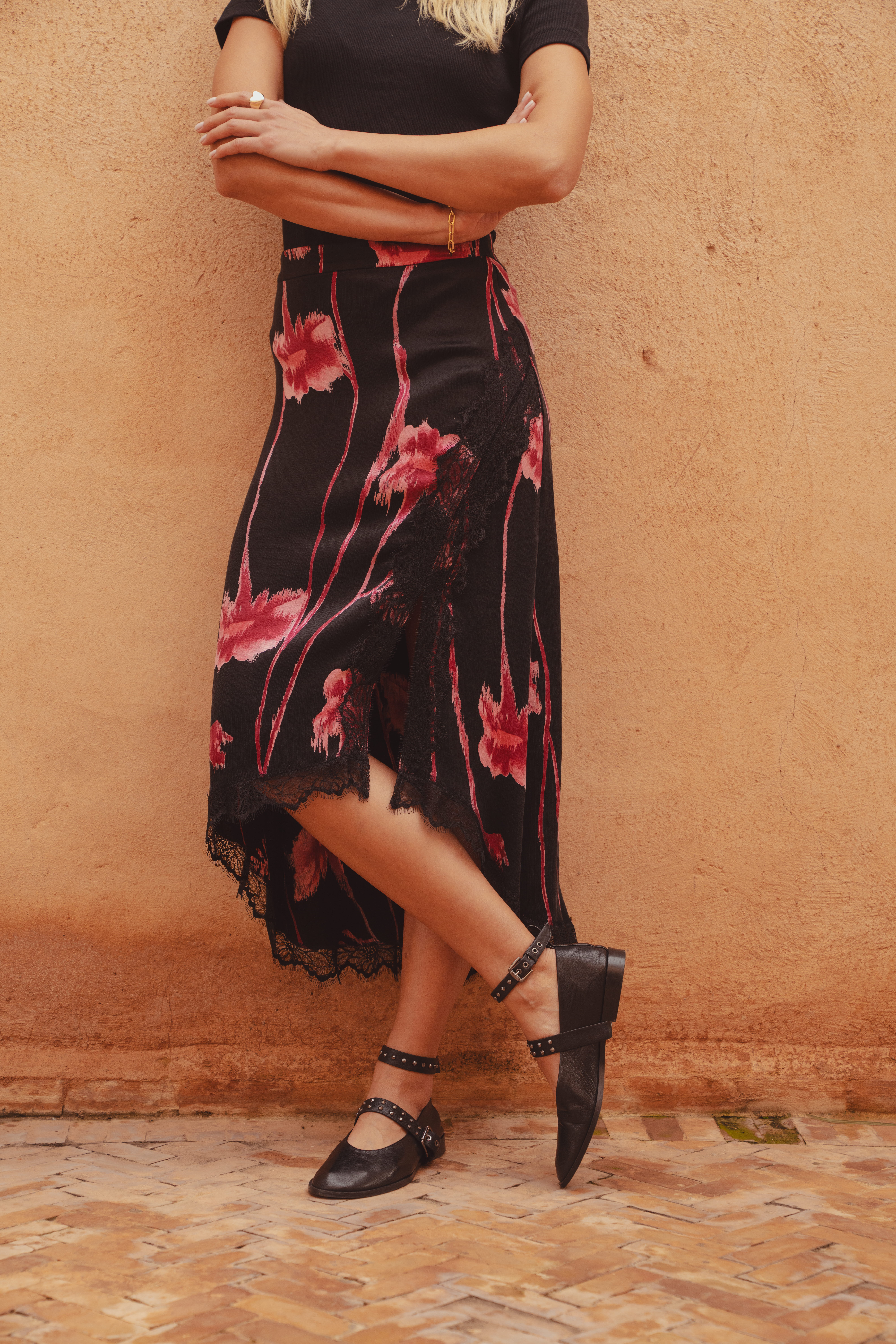A close up shot of the Harper Midi Skirt by Saint and Sofia. A mid rise style with a side split and lace detailing. It's a floral print skirt with a black background and bright pink flowers. Styled here with leather ballerina flats.