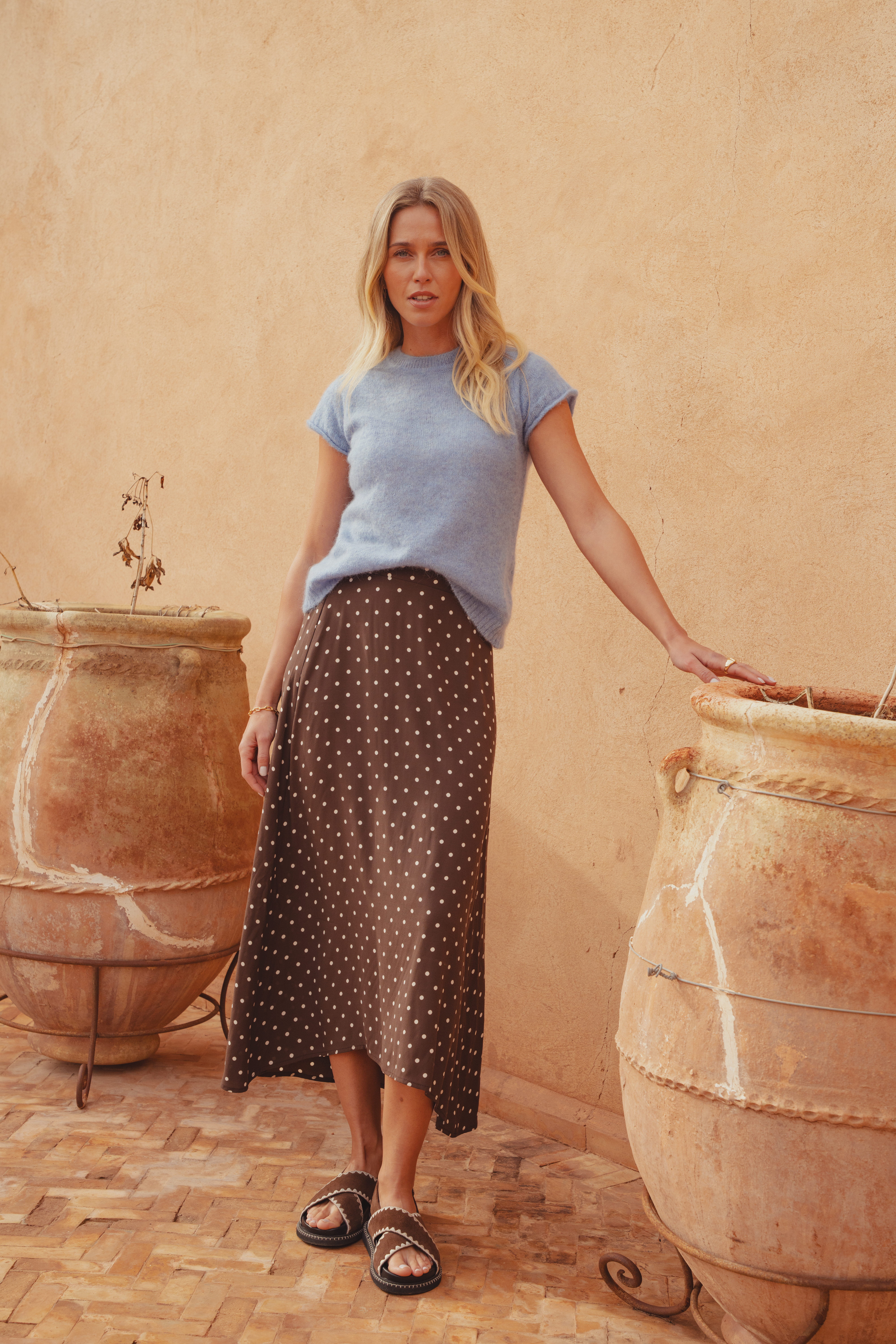 A model standing in front of a clay wall wearing a baby blue knitted top with short sleeves and a crew neckline. She styles it here tucked into a brown and white polka dot midi skirt with cross over brown suede sliders.