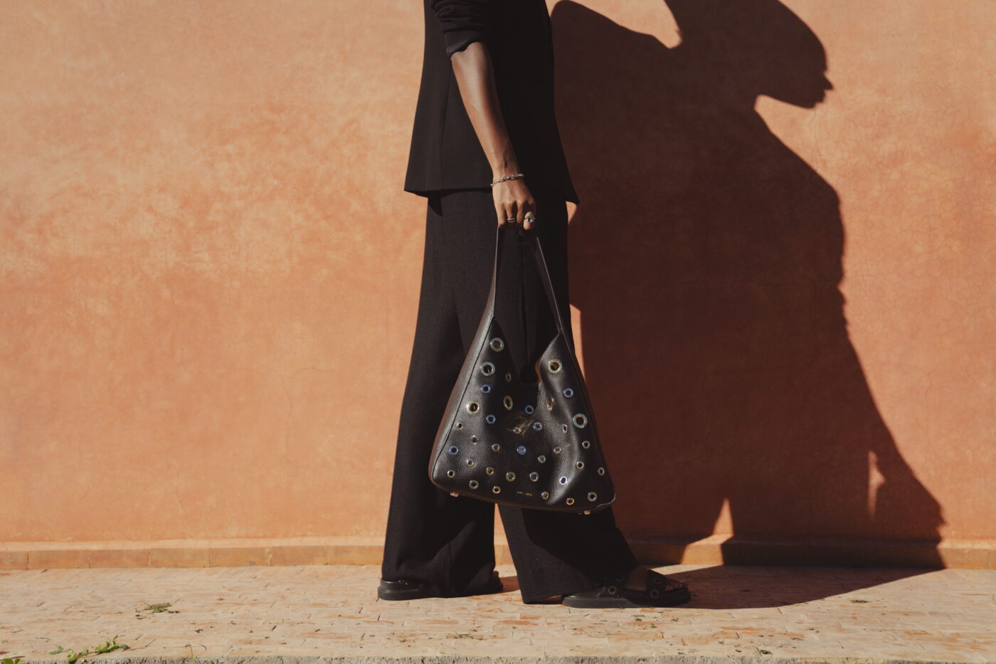 A low down shot of a model walking beside a wall. Her shadow is enlarged beside her. She wears a black blazer, wide-leg black trousers, and in her hand is the black Allana Eyelet bag by Saint and Sofia. A leather hobo-style bag with silver and gold eyelet details.
