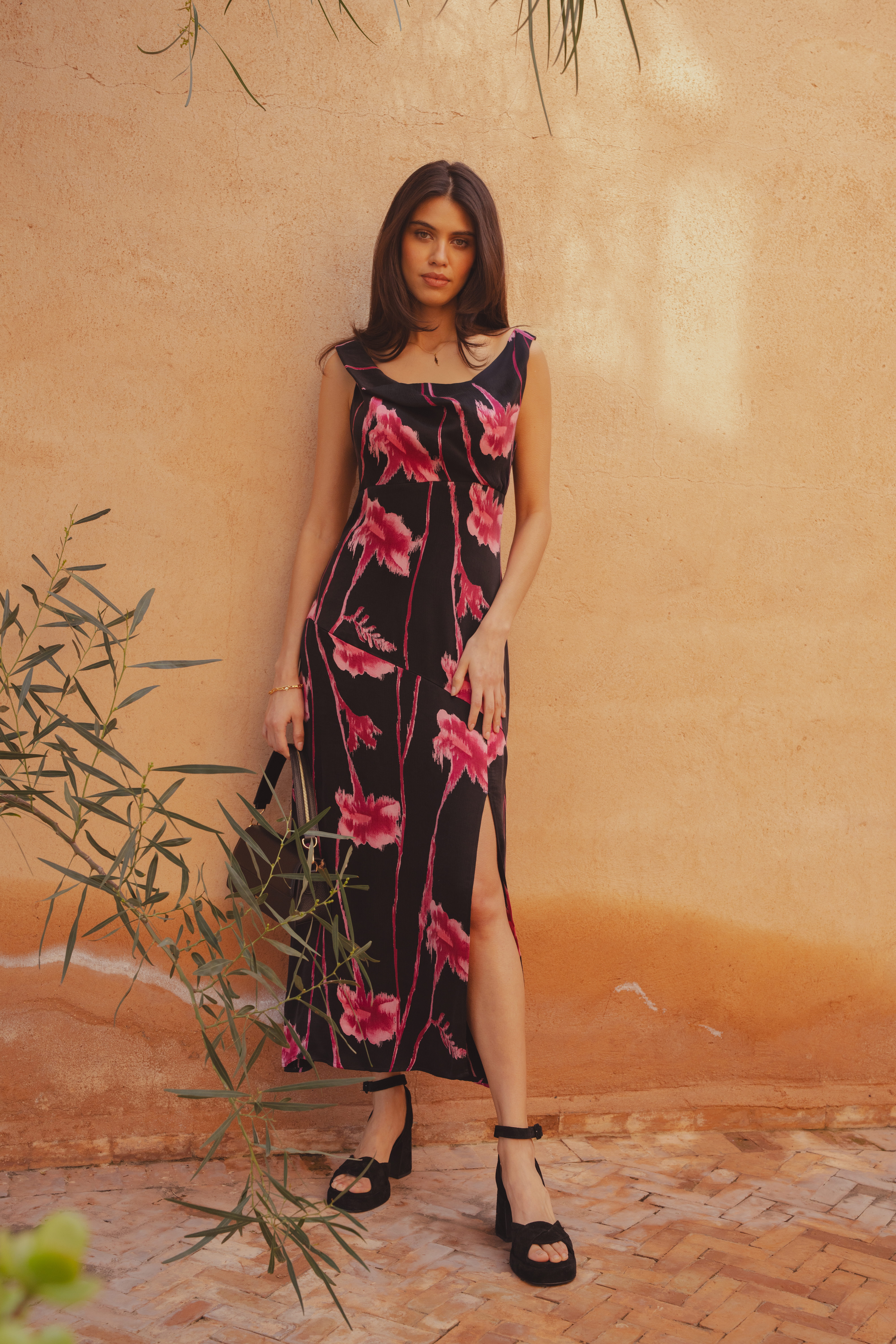 A model wearing the Alexis Dress by Saint and Sofia. A twisted cowl neck style with wide straps and a split in the skirt. It features a black background with pink flowers. It's worn here with black heels and a black leather handbag.