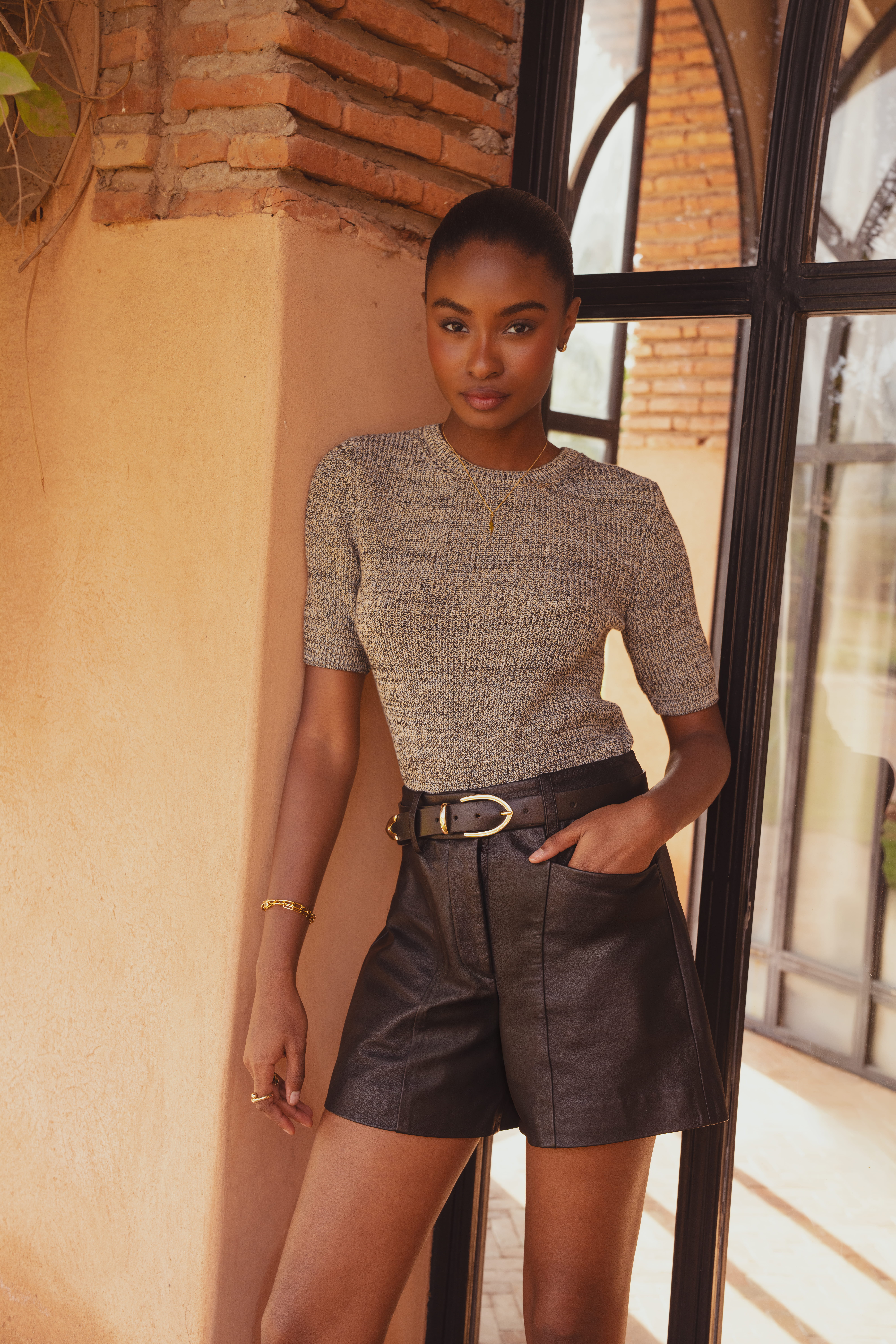 A model leaning against the side of a clay pillar wearing a short sleeve crew neck knitted top in grey from Saint and Sofia with black leather shorts and a black leather belt.