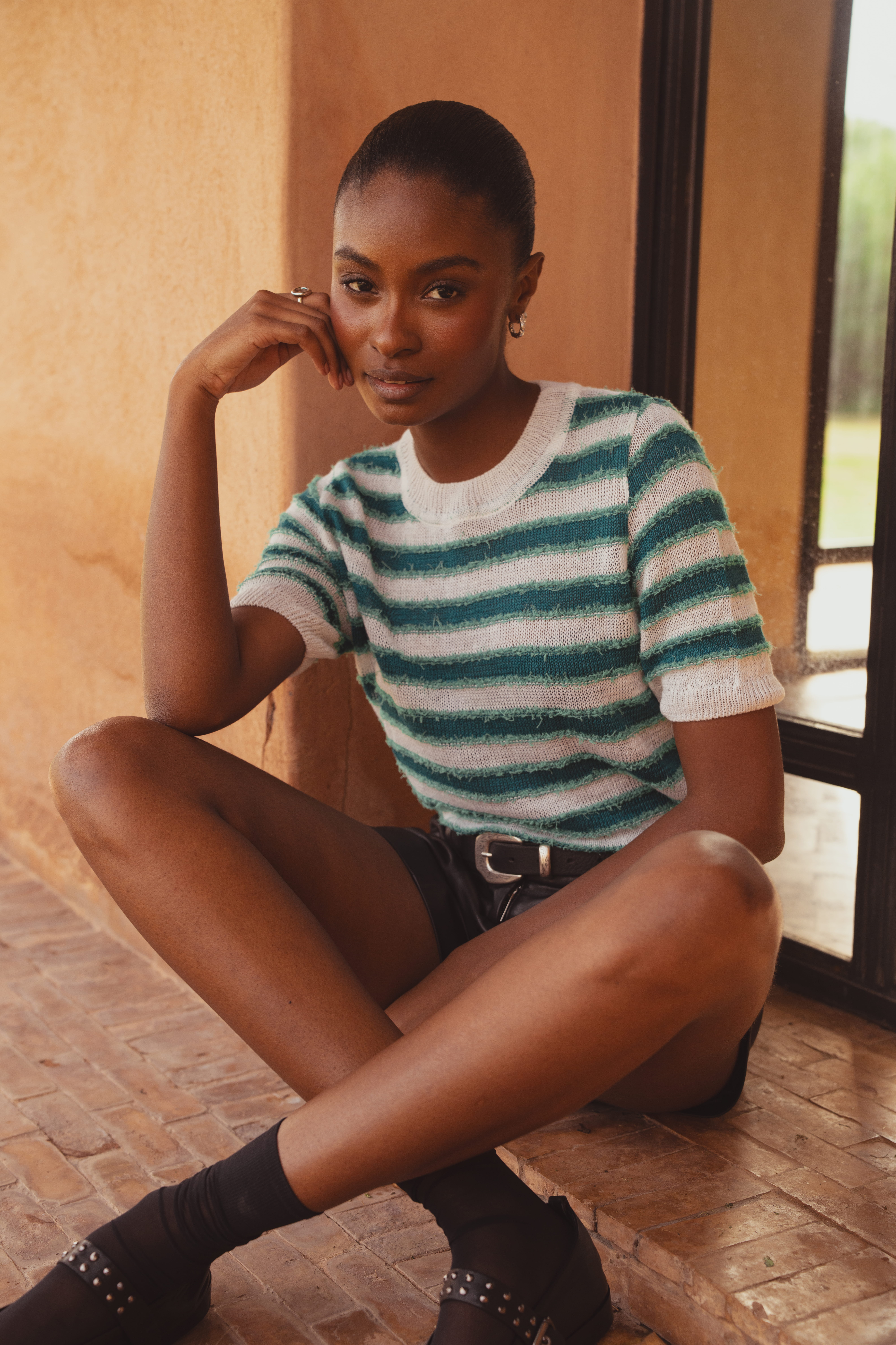 A model sitting cross legged on the ground wearing a green and white striped knitted tee with a crew neckline, short sleeves, and a brushed textured finish. This Fearne Knit Style is by Saint and Sofia.