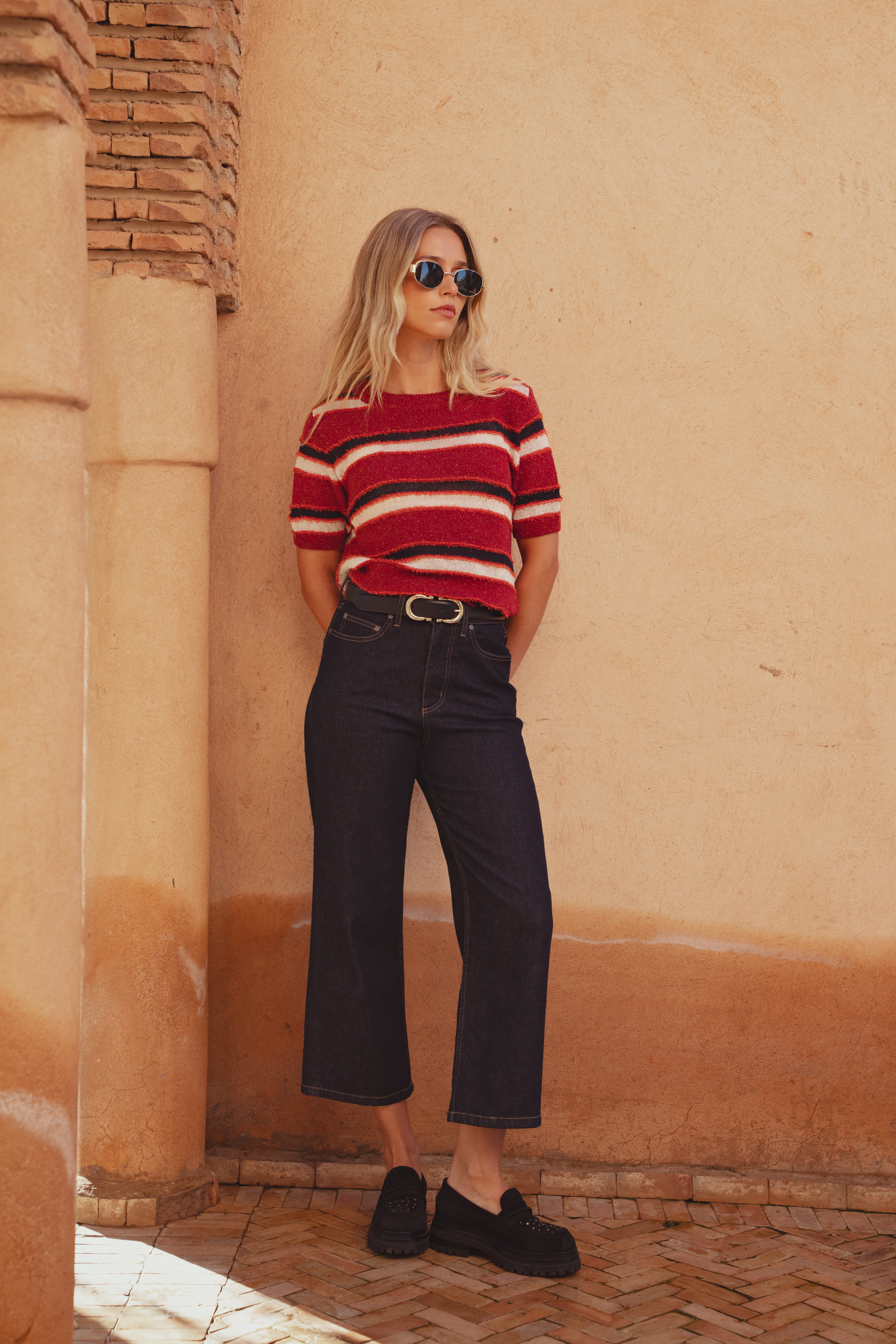 A model standing in front of a clay wall wearing a red, white, and black knitted top with a crew neckline. It's styled here tucked into a pair of cropped denim jeans with loafers.
