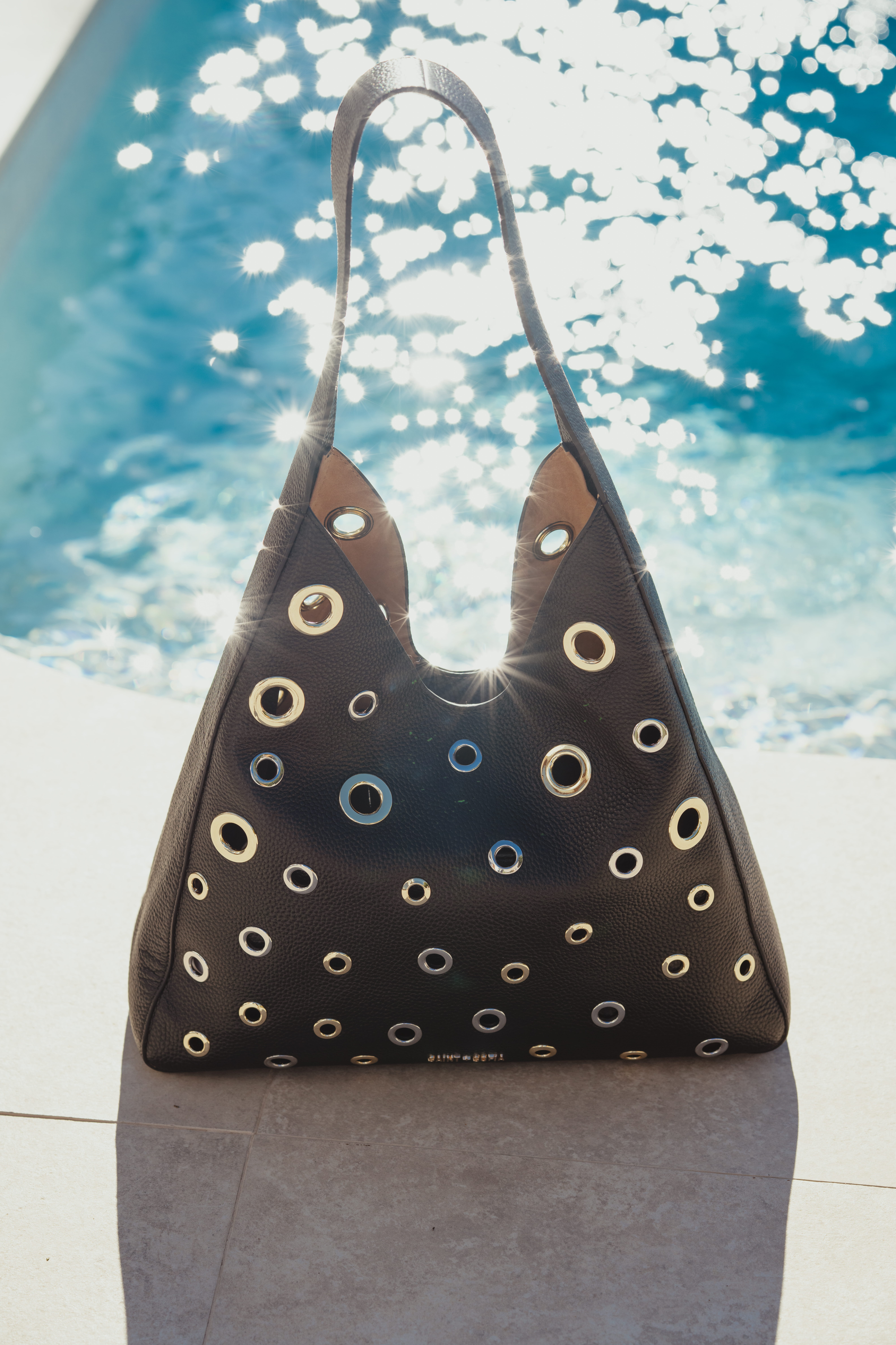 The Allana Eyelet Bag by Saint and Sofia. A leather hobo shaped bag with gold and silver metal eyelet detail and a single thick shoulder strap. Photographed here in front of a pool on a sunny day where the sun is shining off of the water and the metallic details on the bag.