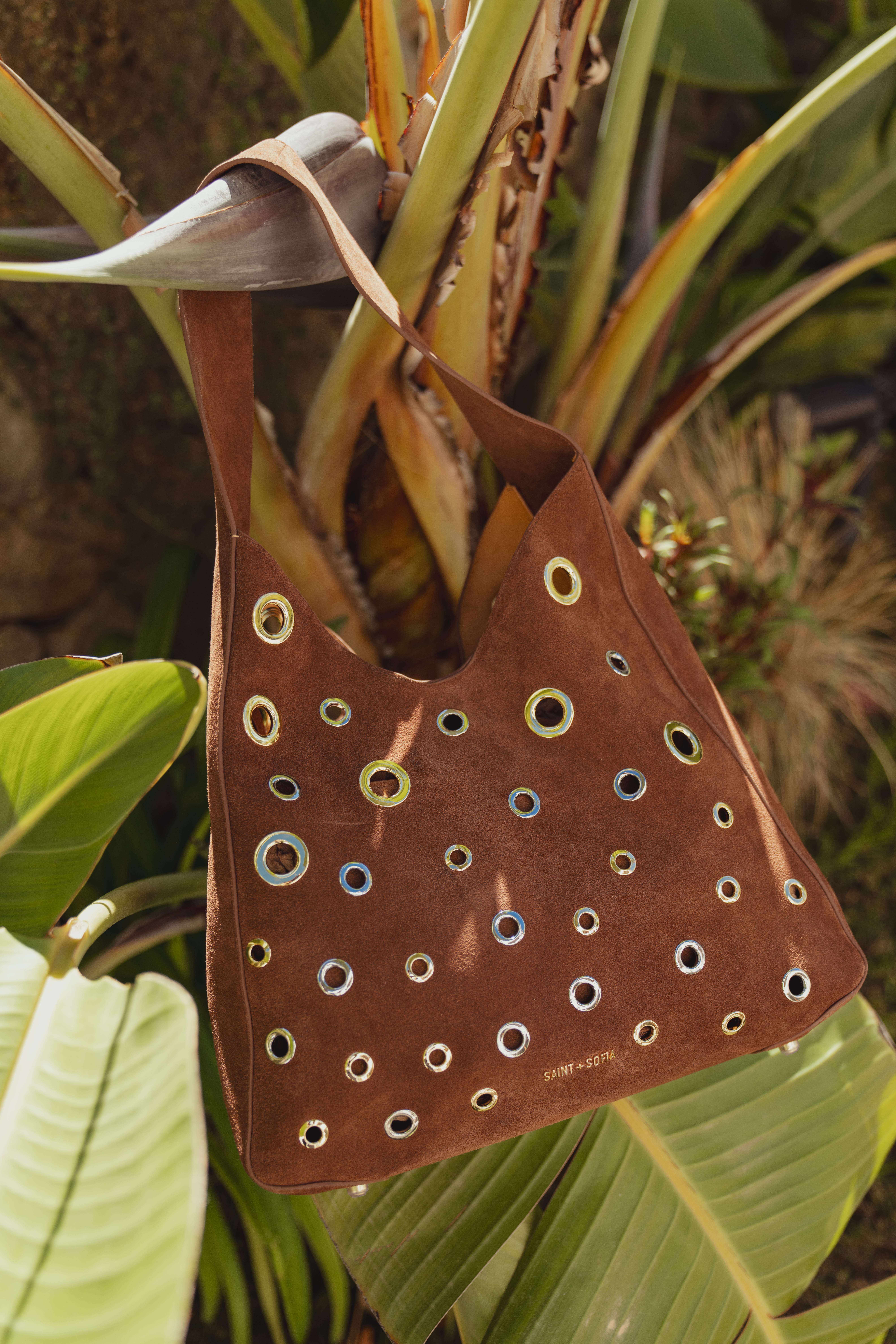 The Allana Eyelet Bag in a a brown suede finish with silver and gold eyelet details across the front and a thick metal shoulder strap. Photographed here hanging from a cactus-like plant.