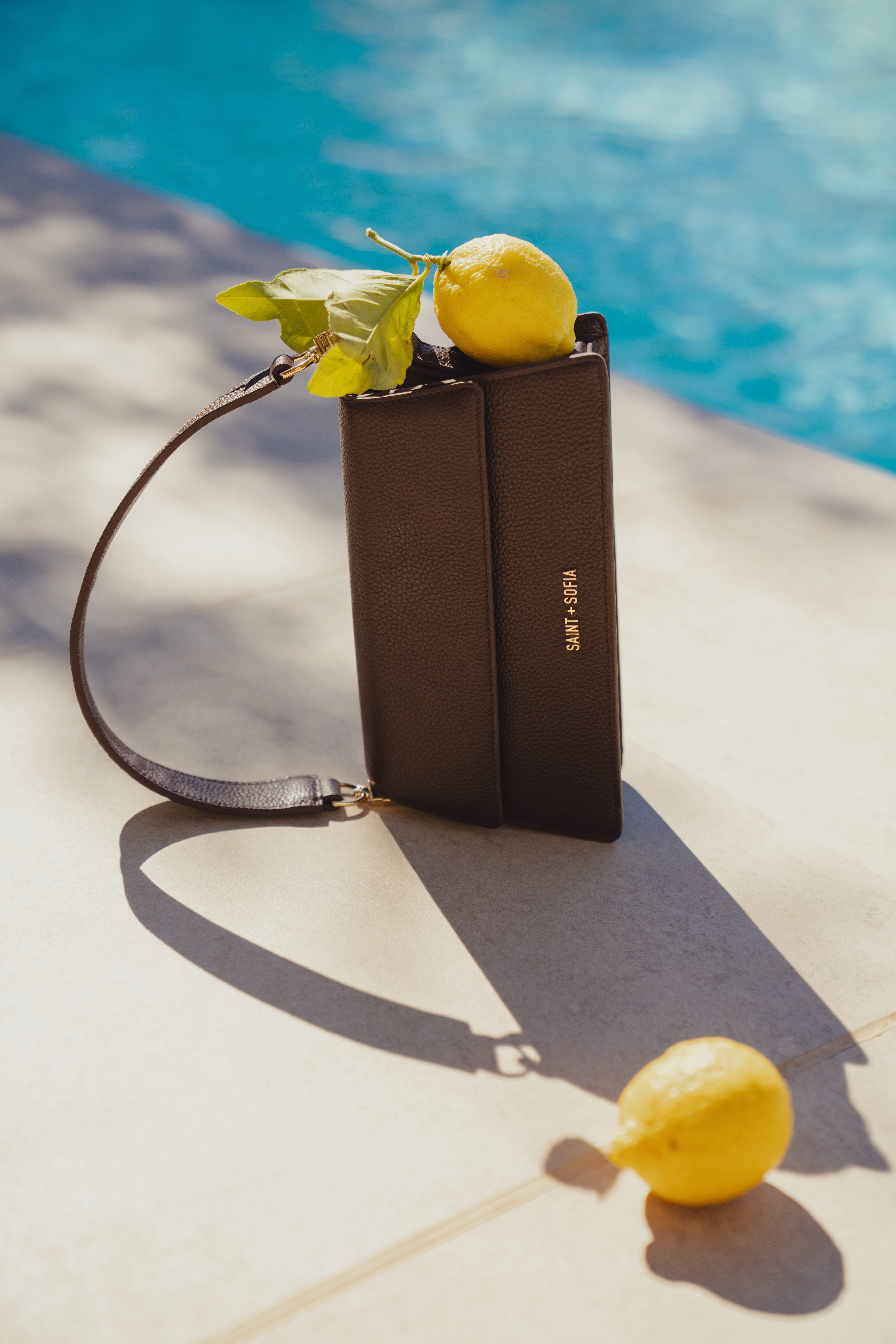 The black Lennox Bag by Saint and Sofia, a miniature trapeze shaped shoulder bag with a branded plaque at the front. Photographed here on its side by a pool with lemons scattered around it.